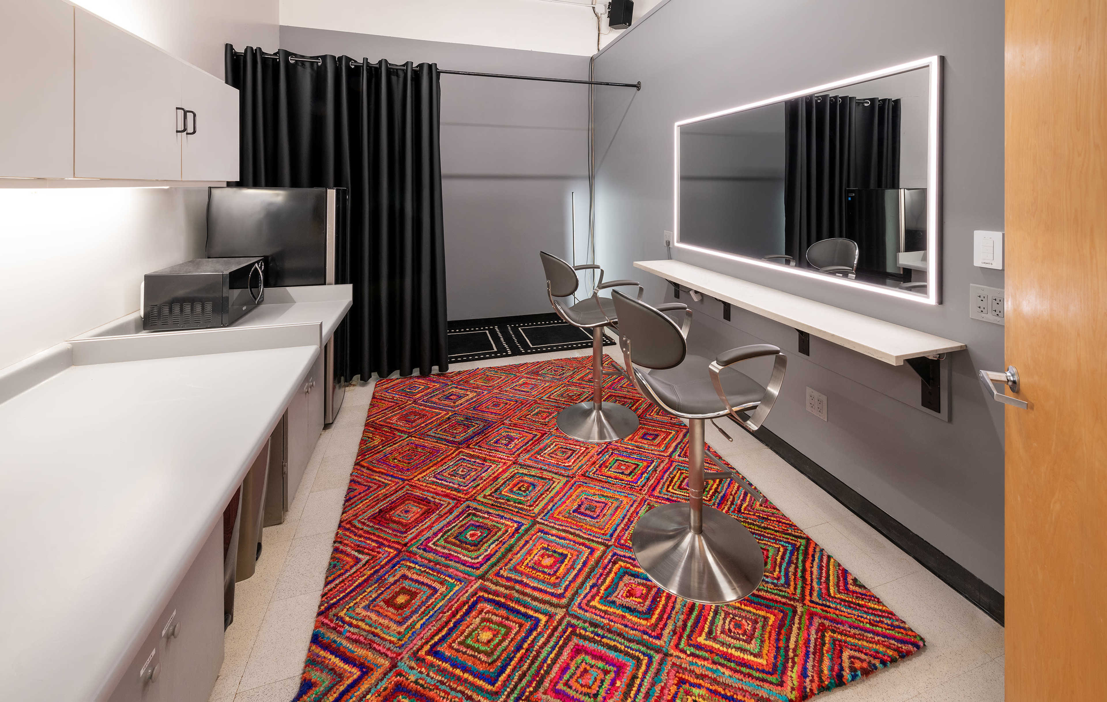A modern makeup room featuring two high stools, a backlit mirror, and a colorful geometric rug on the floor.