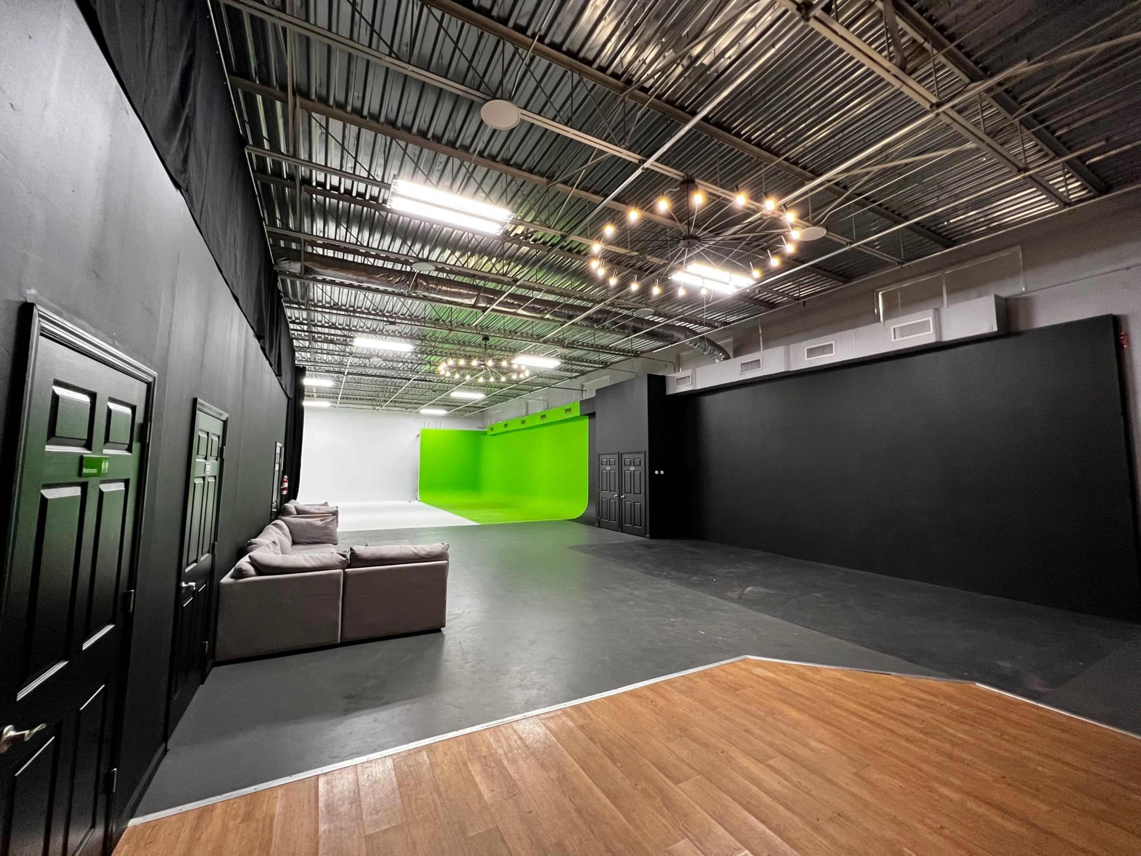 A spacious studio with gray flooring and black walls, featuring a green screen area at one end and a seating area with a couch.