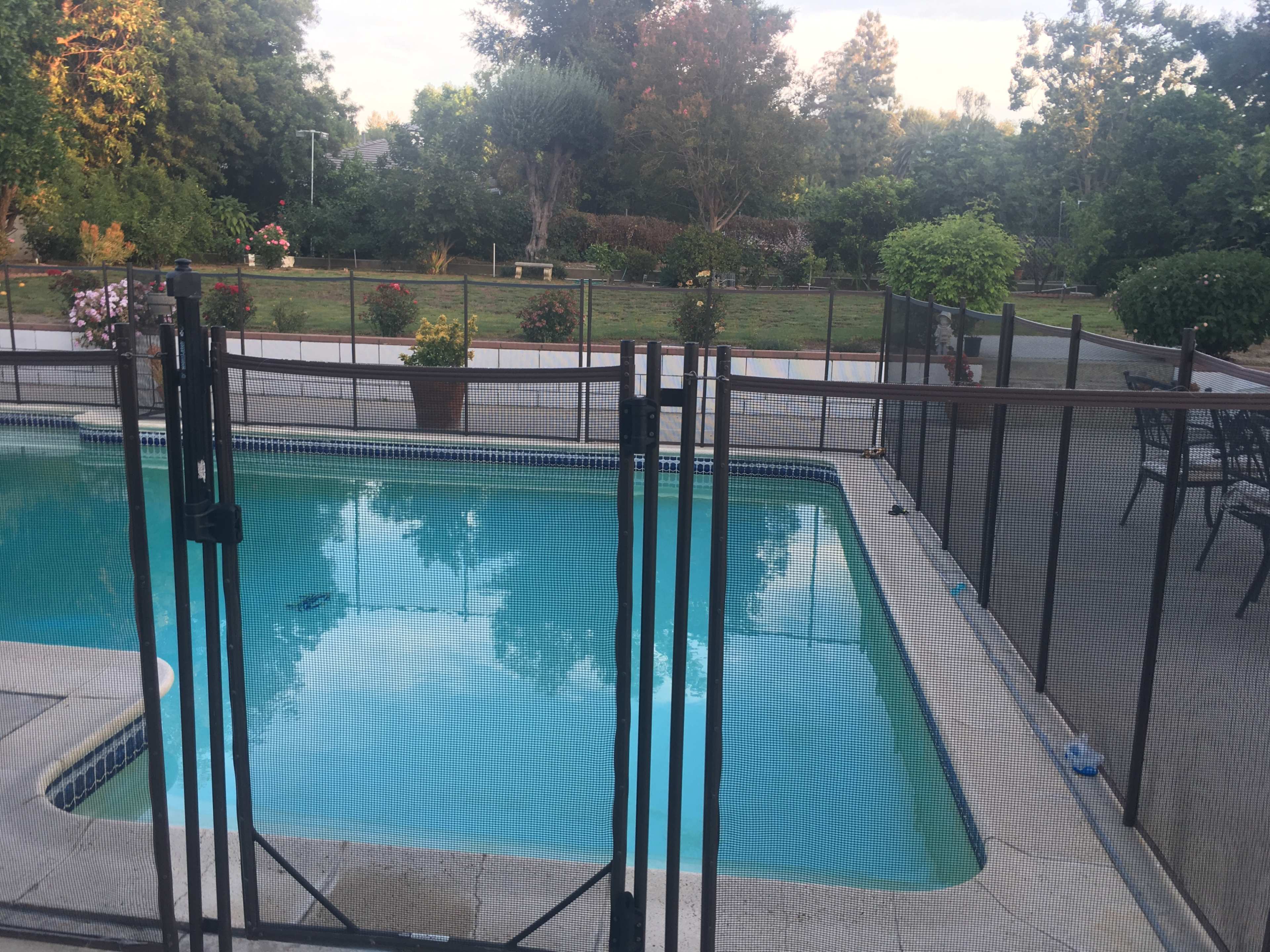 The image shows a swimming pool surrounded by a safety fence, with a landscaped garden and seating area visible in the background.