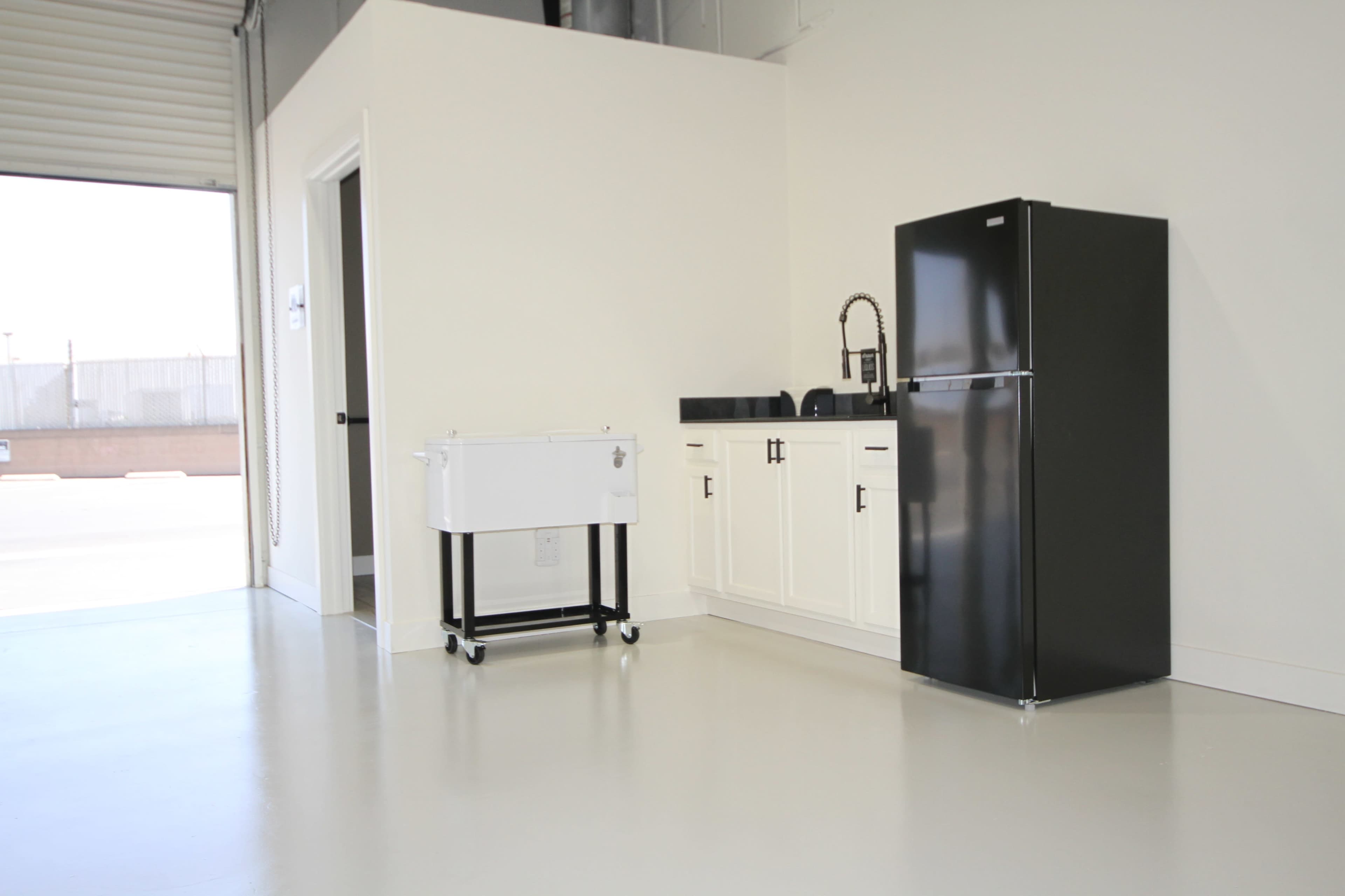 The image shows a minimalist kitchen area featuring a black refrigerator, a sink with a faucet, and a white cabinet on wheels in a bright, open space.