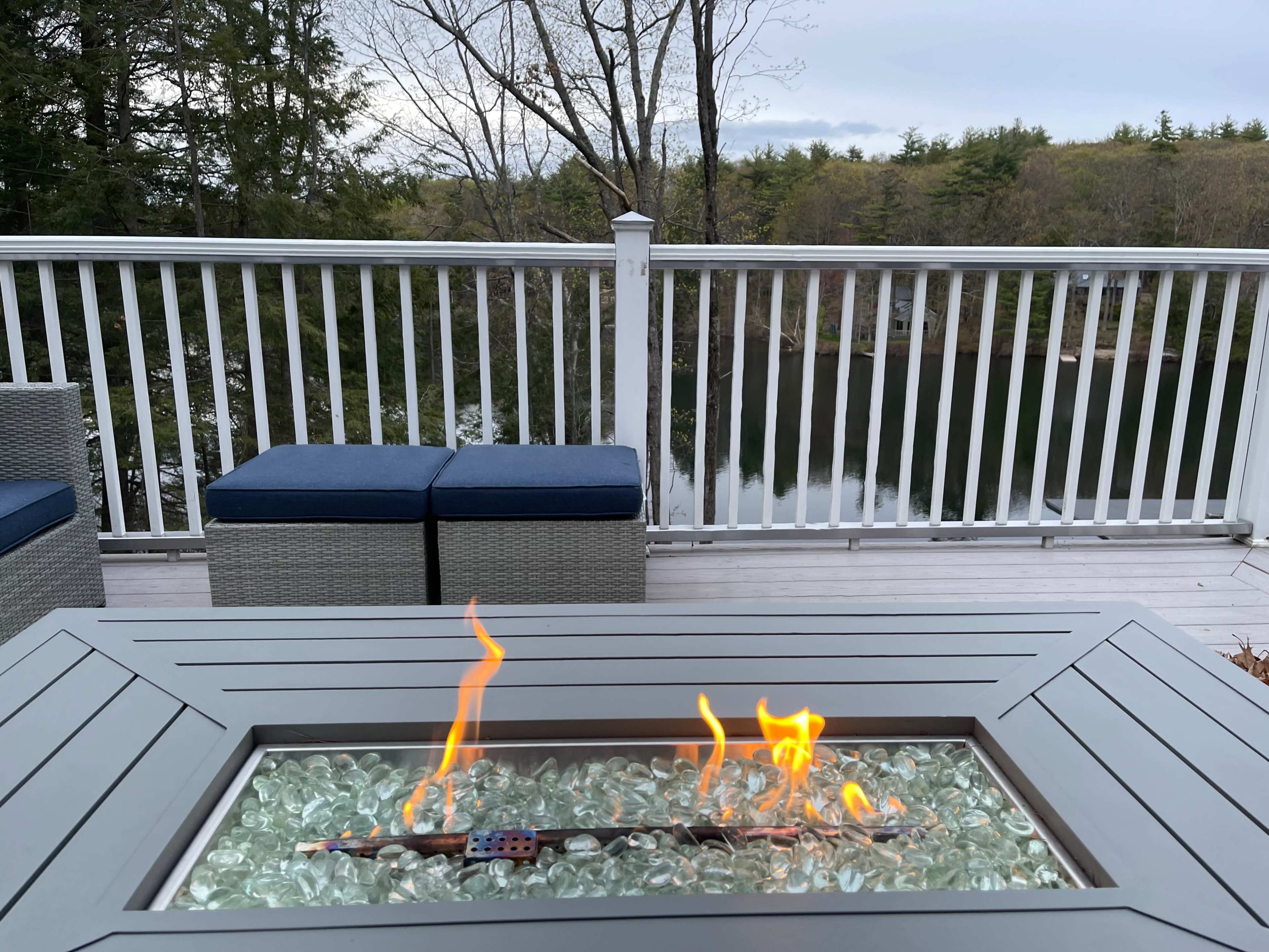 A flame flickers in a fire pit surrounded by a wooden deck and a view of a reflective lake beyond.