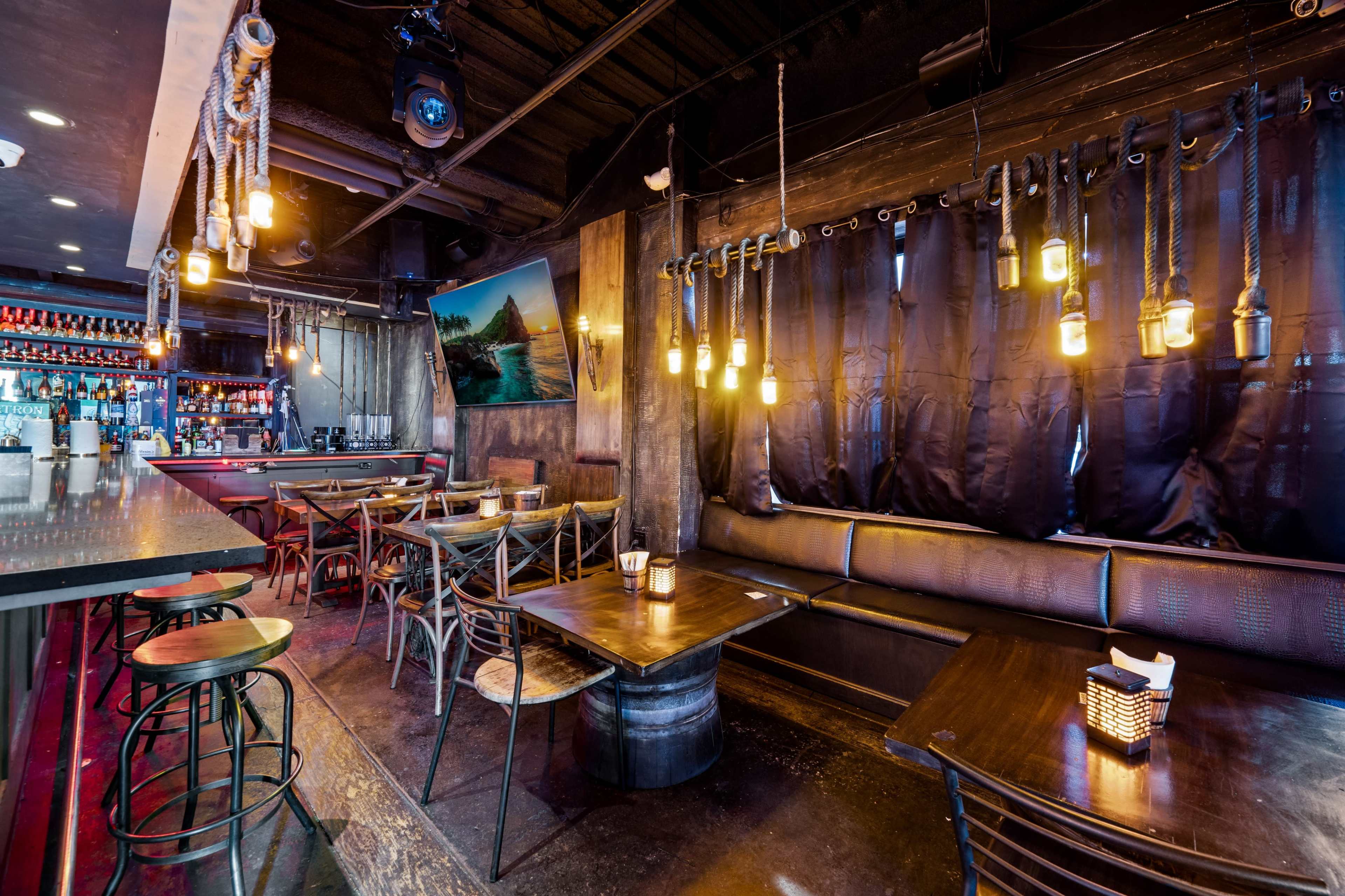 A darkly lit bar interior with wooden tables, high stools, and decorative pendant lights hanging from the ceiling.