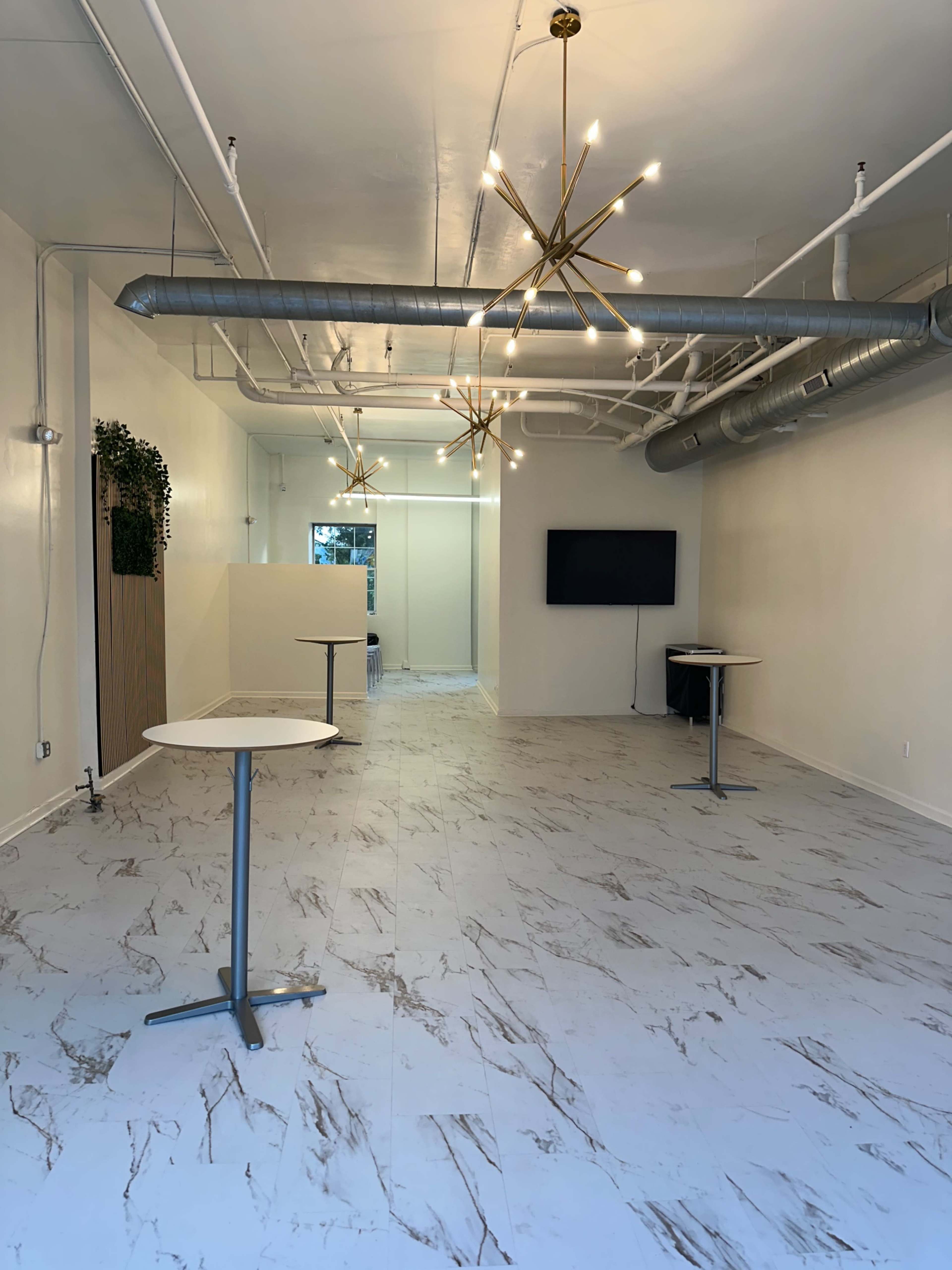 The image shows a spacious, well-lit interior with modern lighting fixtures, featuring two round tables and a television mounted on the wall.