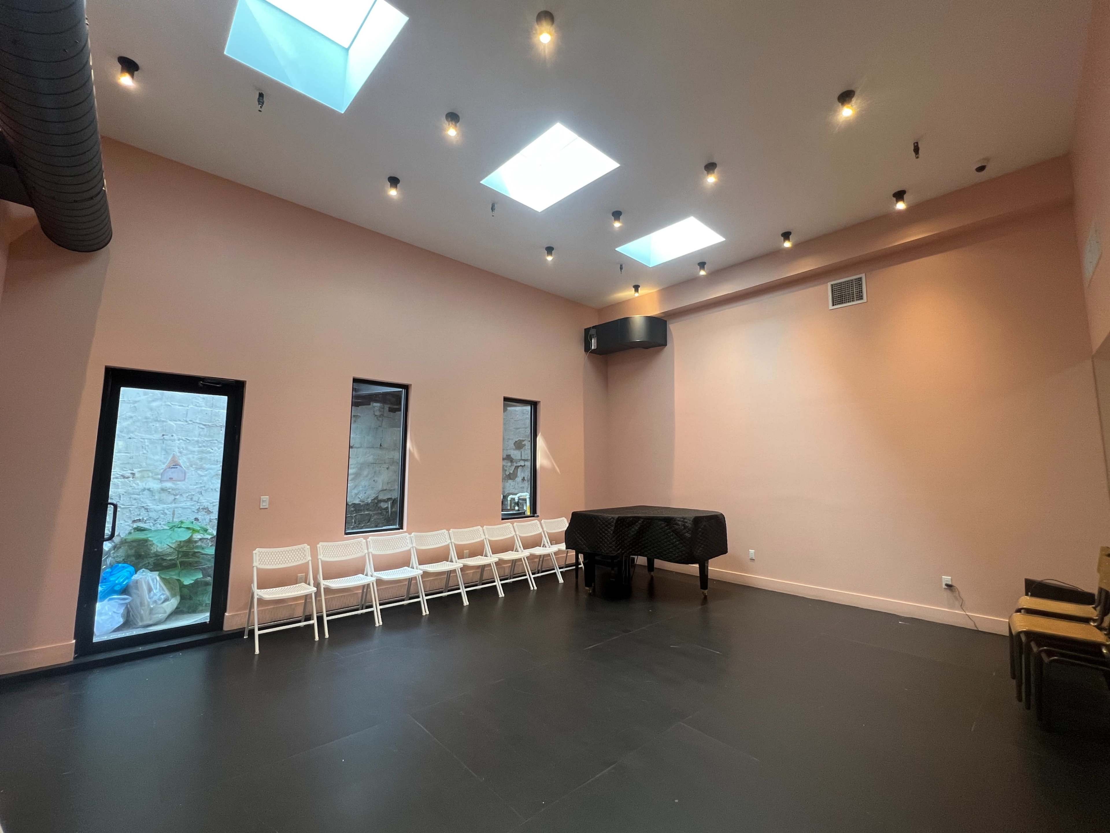 The image shows a spacious room with pink walls, a black table in the center, and several white chairs lined up against one wall, illuminated by ceiling lights and skylights.
