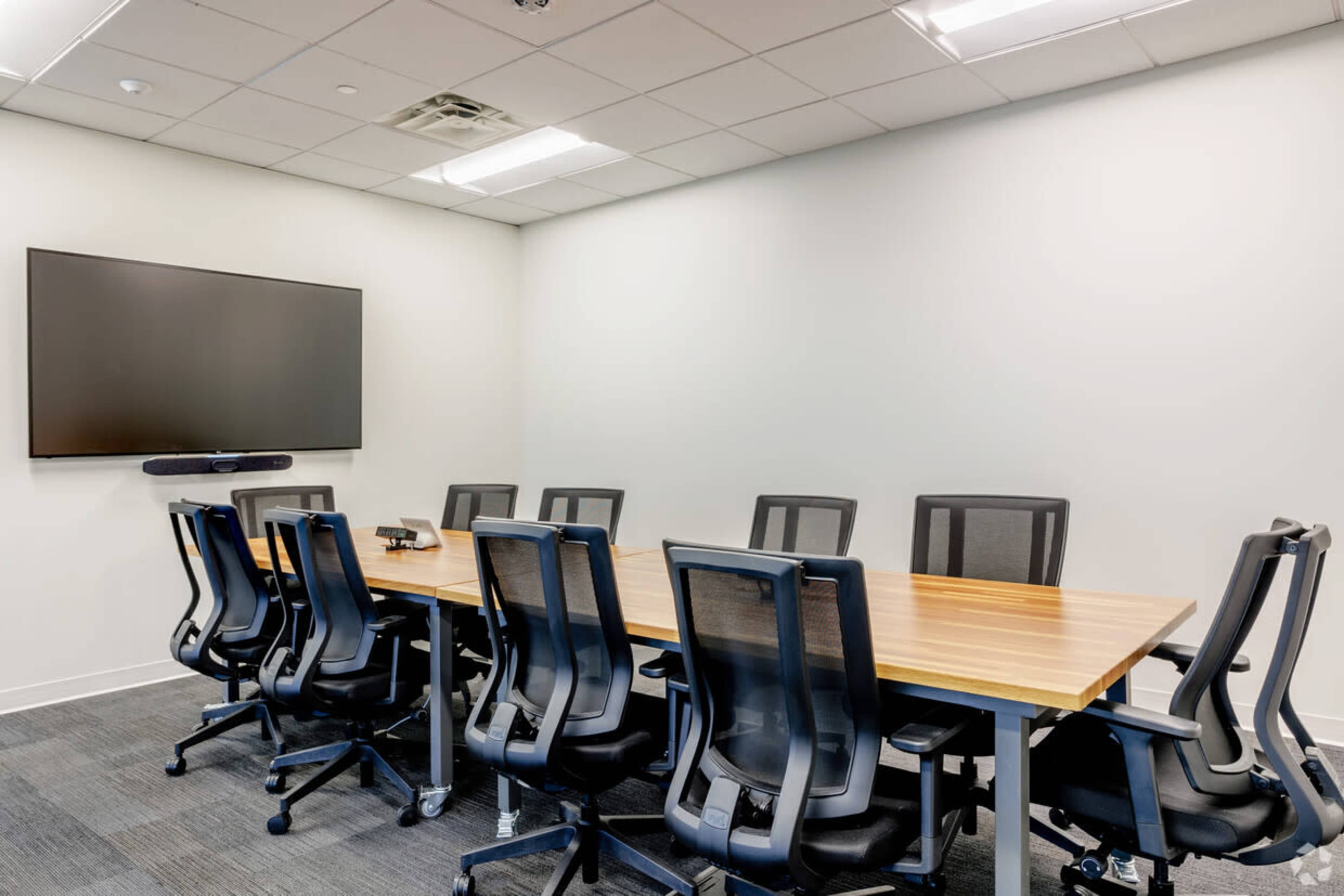 A sparsely furnished meeting room features a long wooden table surrounded by black office chairs and a mounted television on the wall.