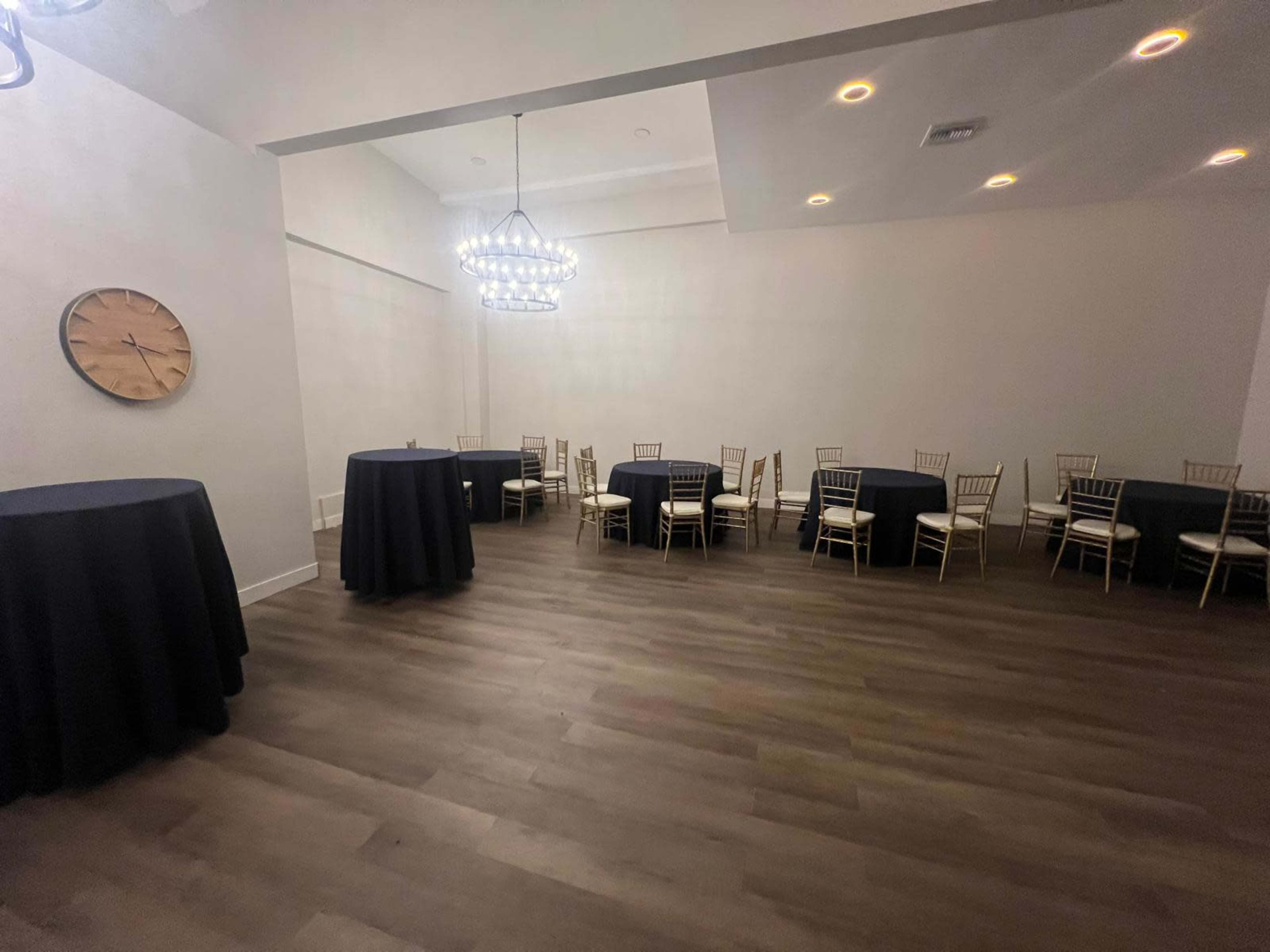 The image shows a spacious, empty event room with round tables covered in dark tablecloths and chairs arranged around them.