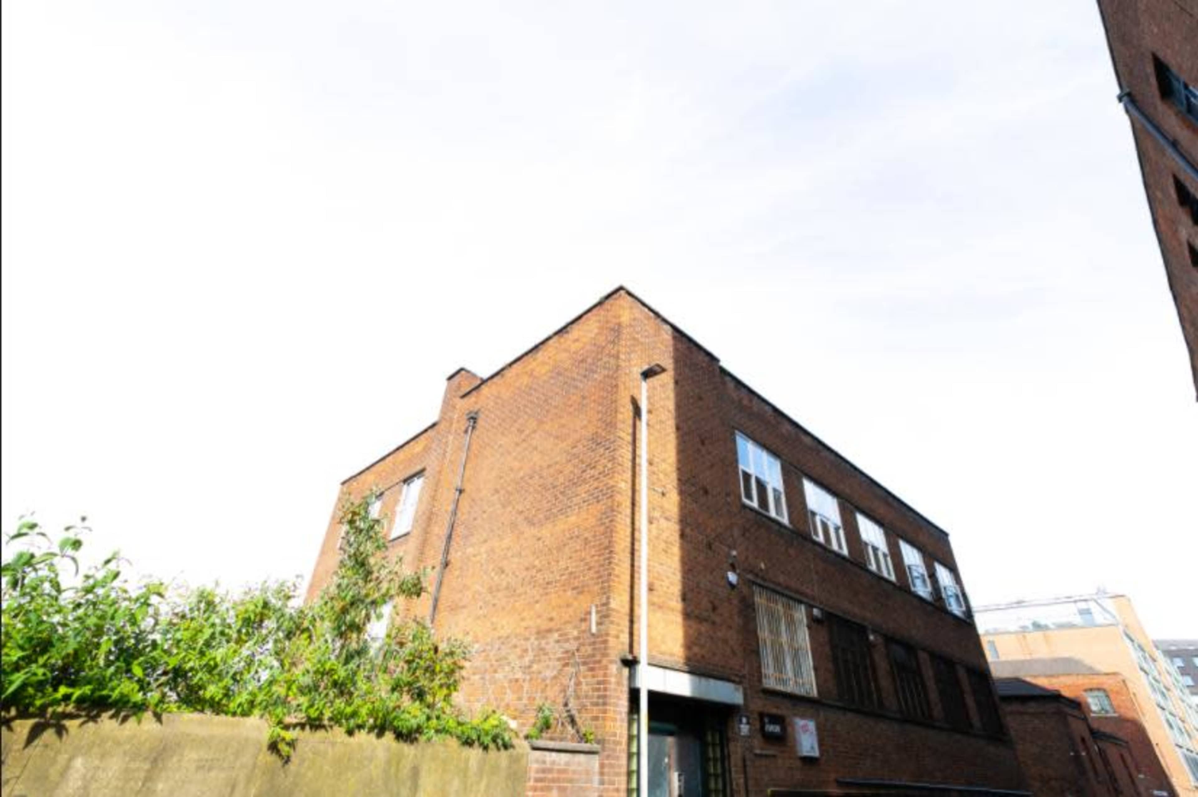 A brick building with a flat roof is seen from a low angle, surrounded by greenery and other structures.