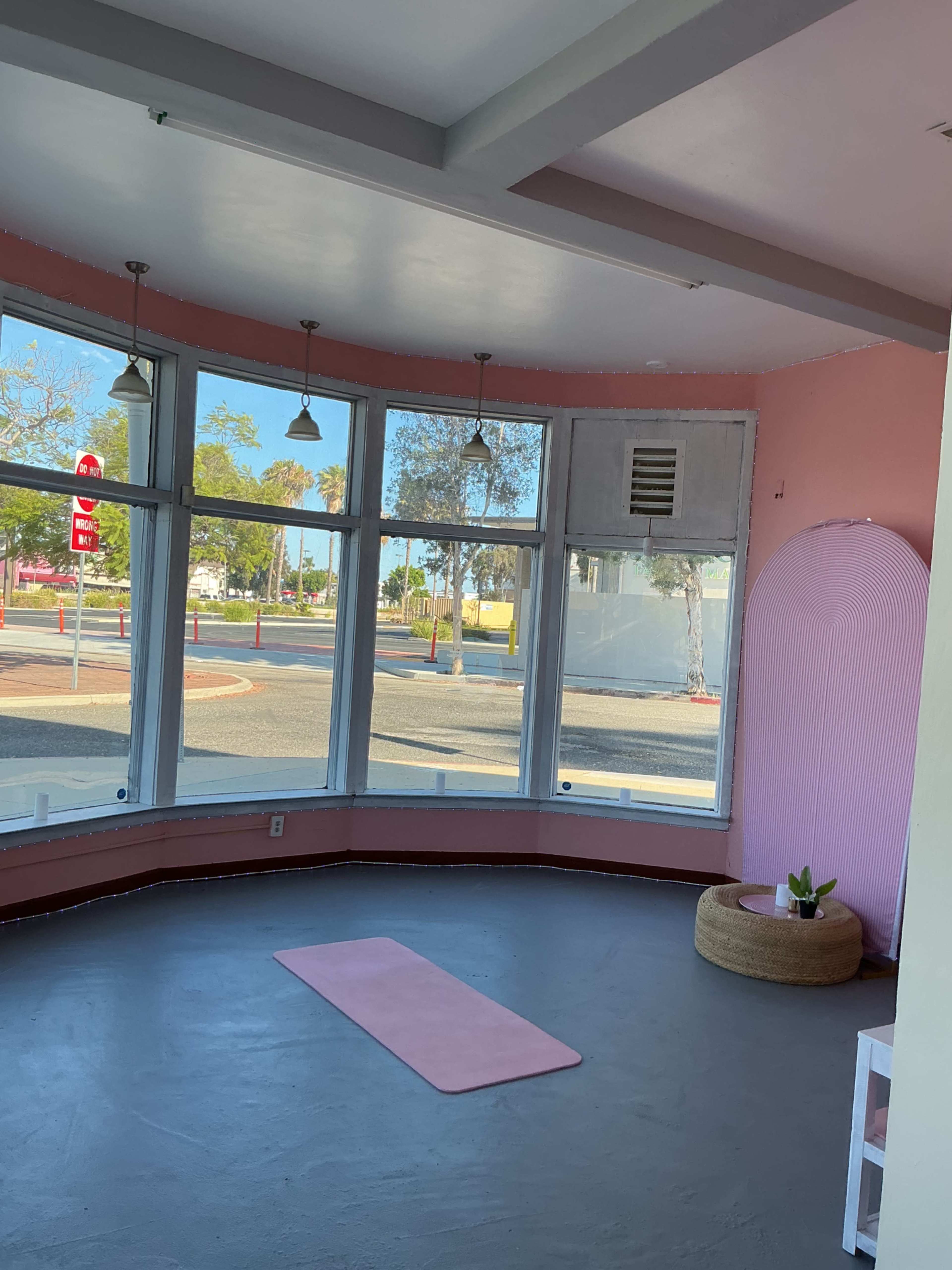 The image shows a bright, modern studio space with large windows, a pink yoga mat on the floor, and a round seating area.