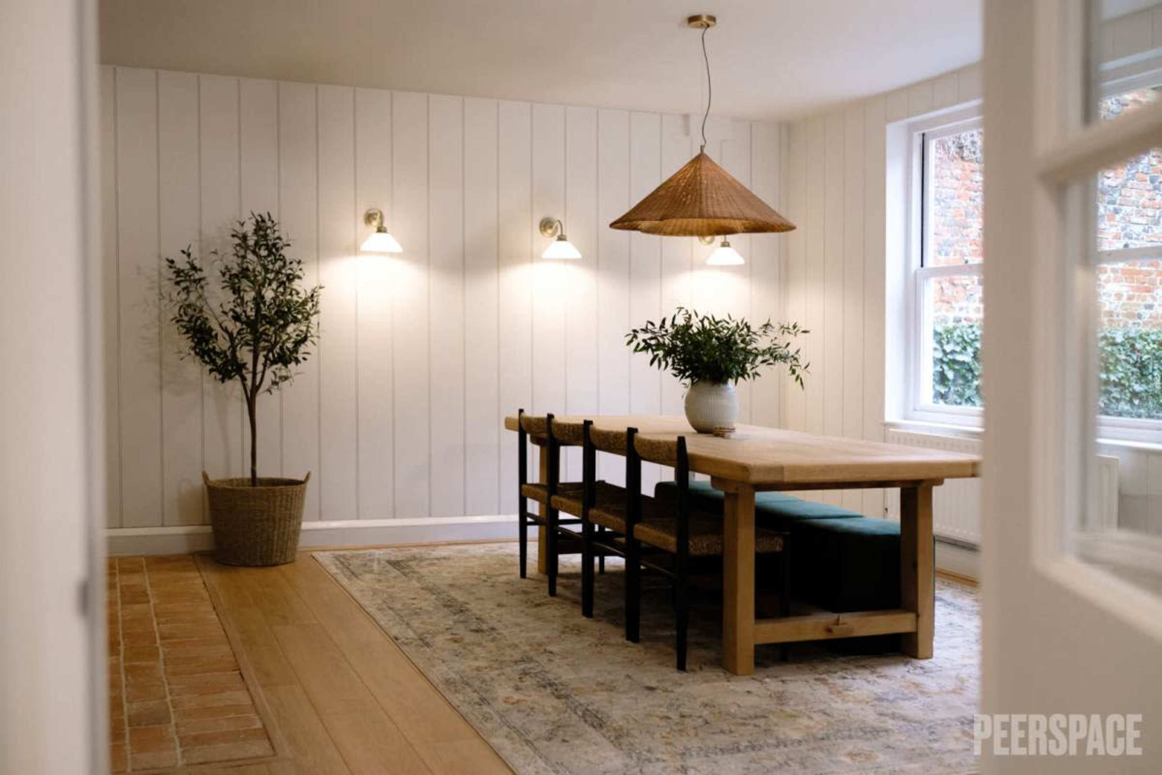 A minimalistic dining area featuring a wooden table with six chairs, a woven pendant light, and a potted plant in a corner.