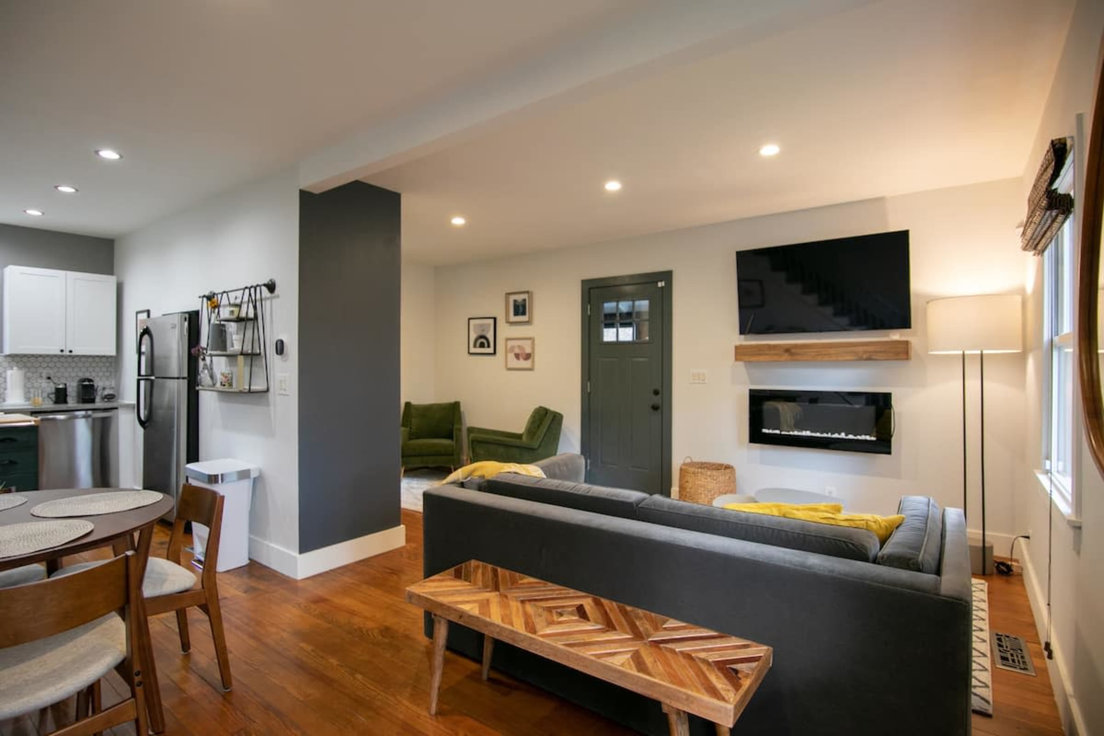 A modern living room with a gray sofa, a wooden coffee table, and a mounted TV on the wall, complemented by a green armchair and a doorway leading outside.
