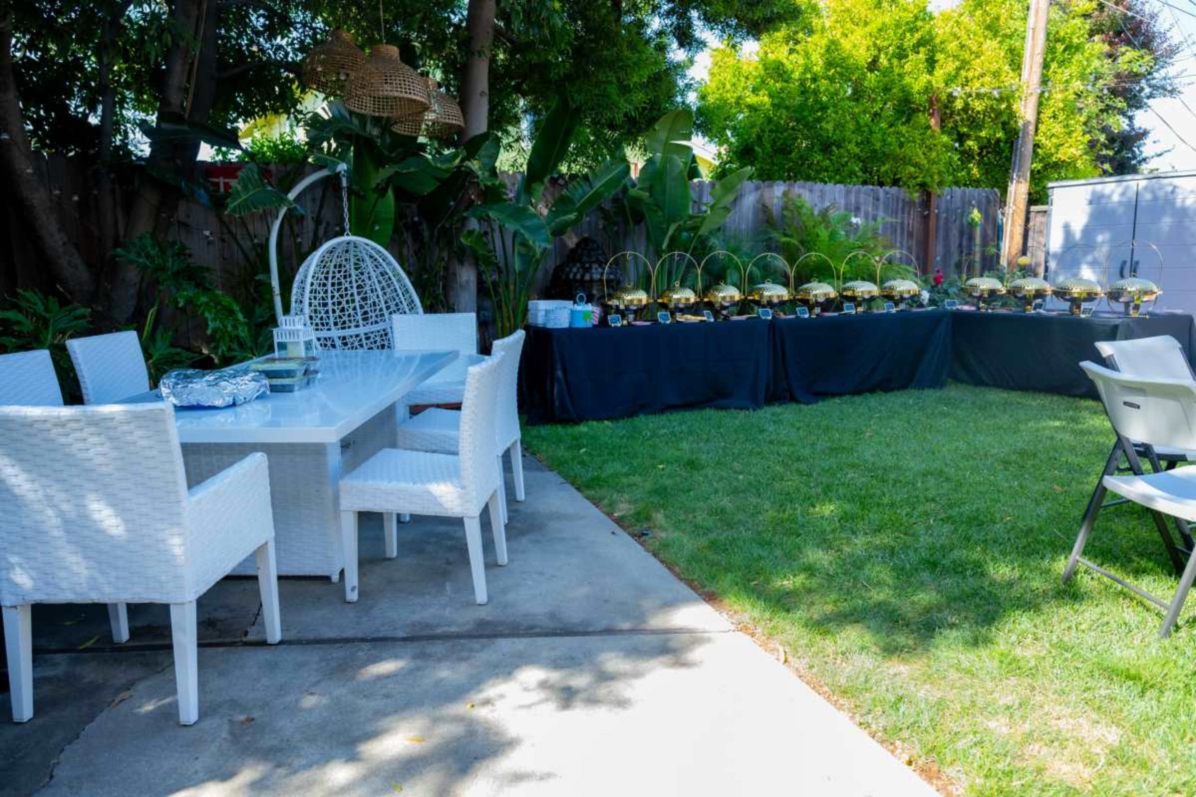 A well-arranged backyard features a dining area with white furniture and a serving table covered with black cloth displaying decorative items amidst green foliage.