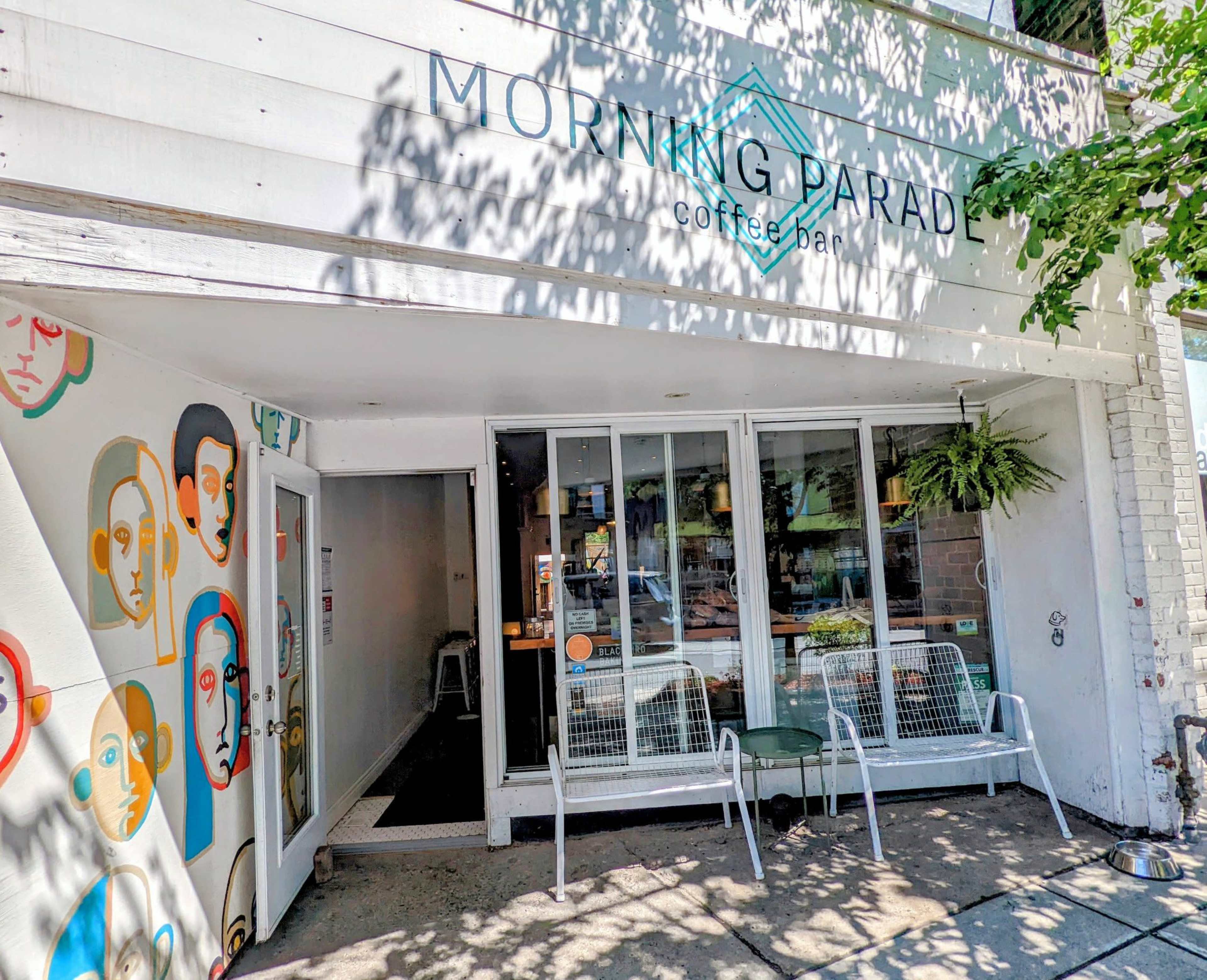 The exterior of a coffee shop named "Morning Parade," featuring a mural of faces on one wall and outdoor seating.