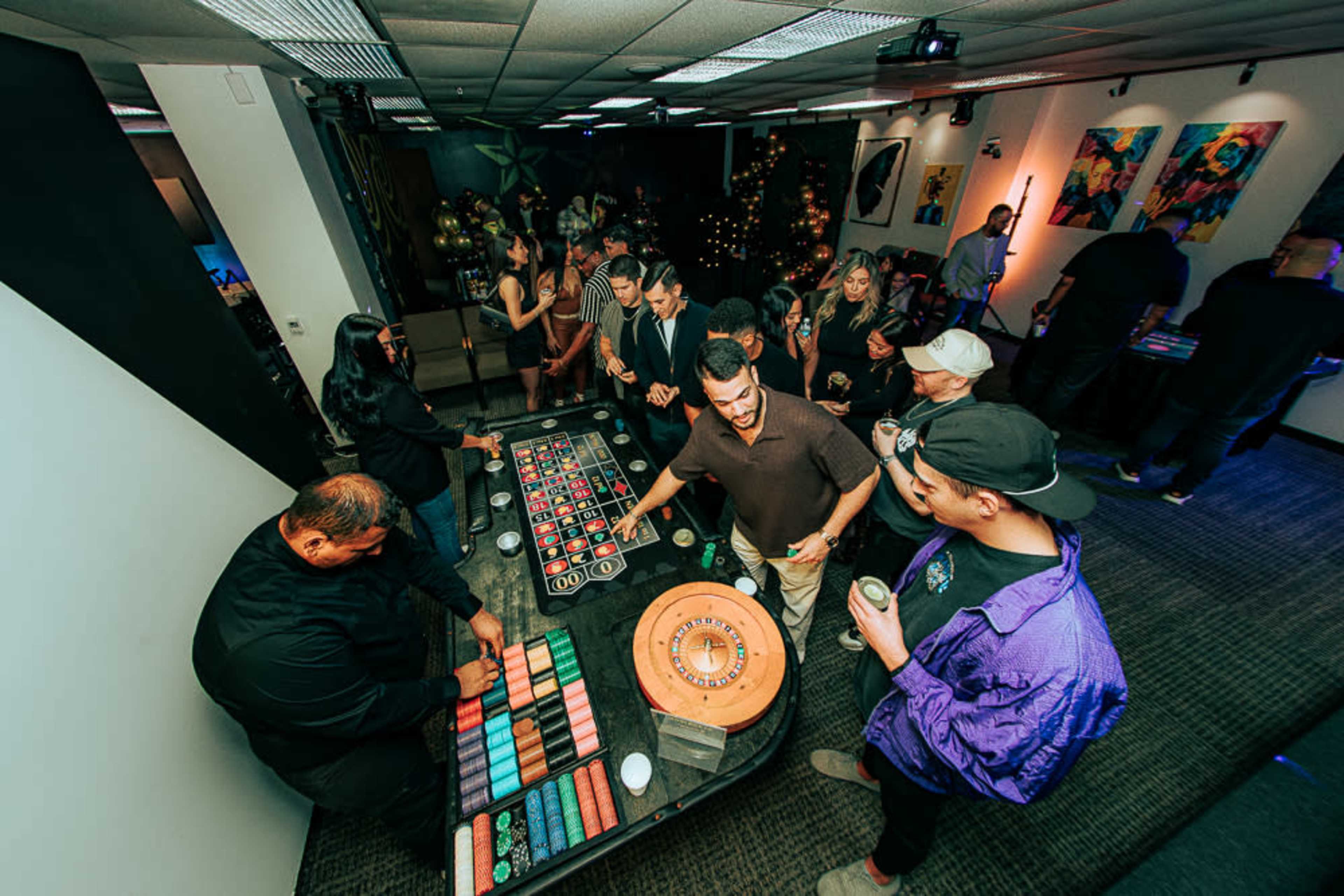 A group of people are gathered around a casino-style roulette table in a lively event space decorated with artwork and lights.