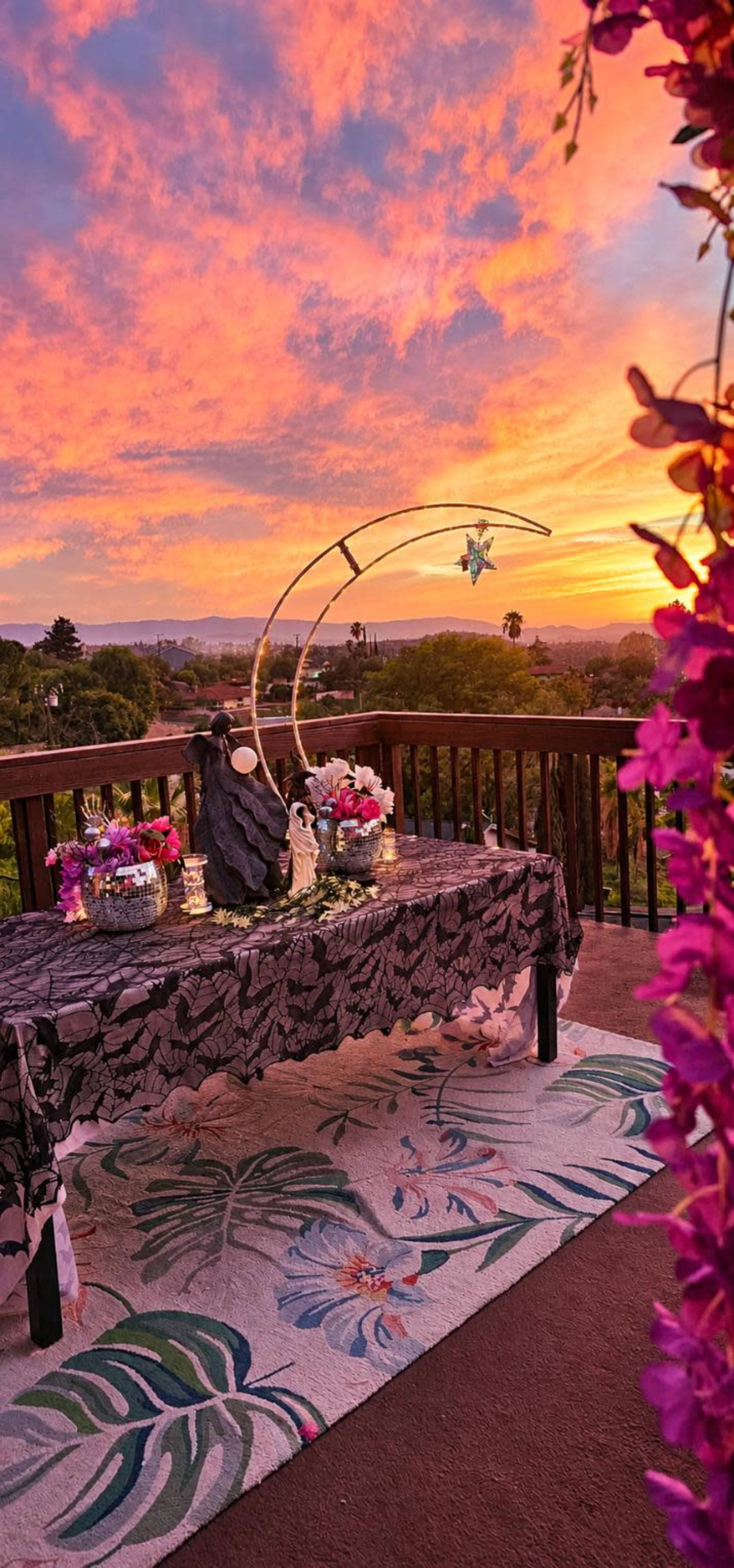 A beautifully arranged outdoor table draped with a black floral tablecloth is set against a vibrant sunset sky, with colorful flowers and decorative items displayed on top.