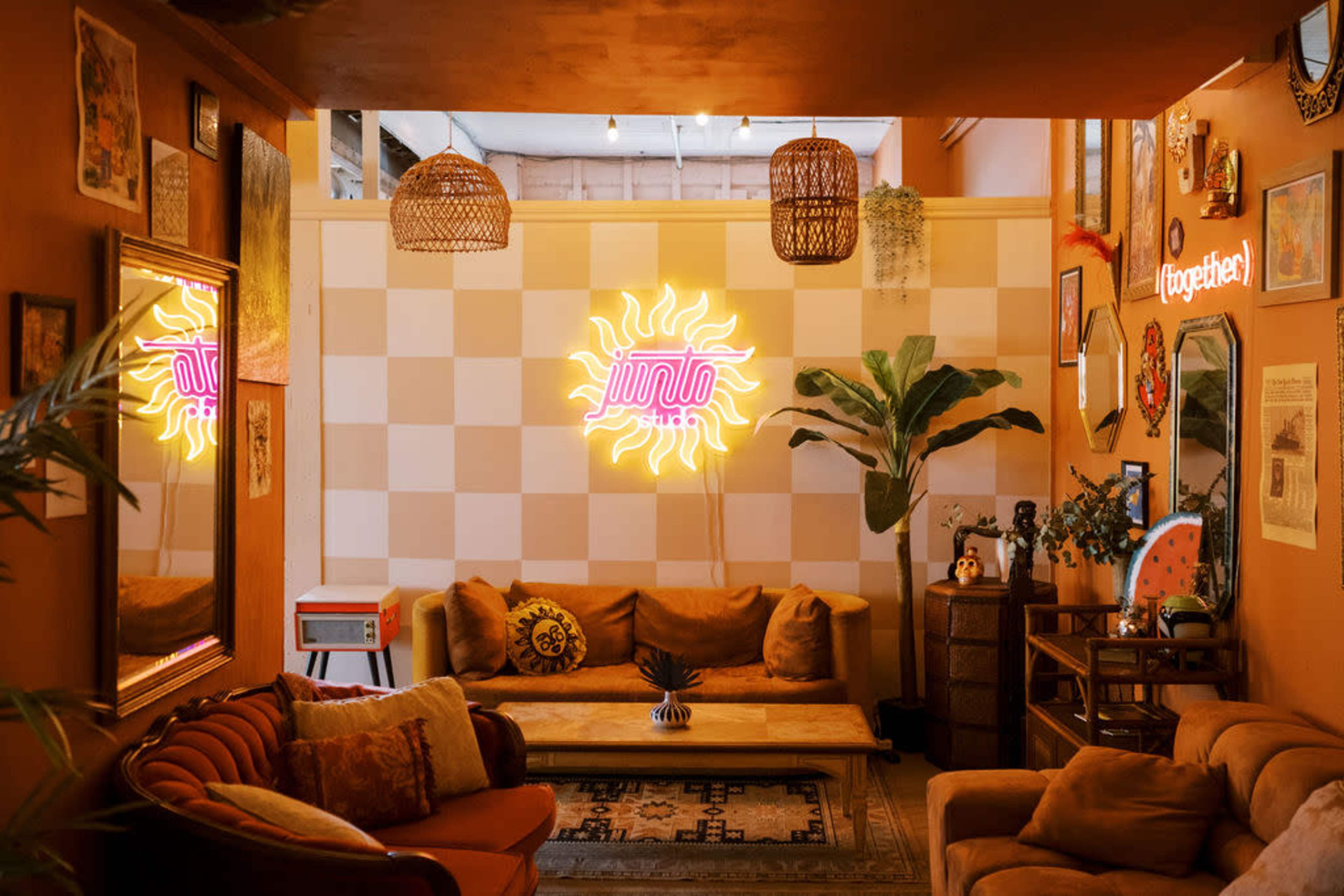 The image shows a cozy living space featuring a neon sun sign, multiple patterned cushions on a sofa, and decorative plants on the walls.
