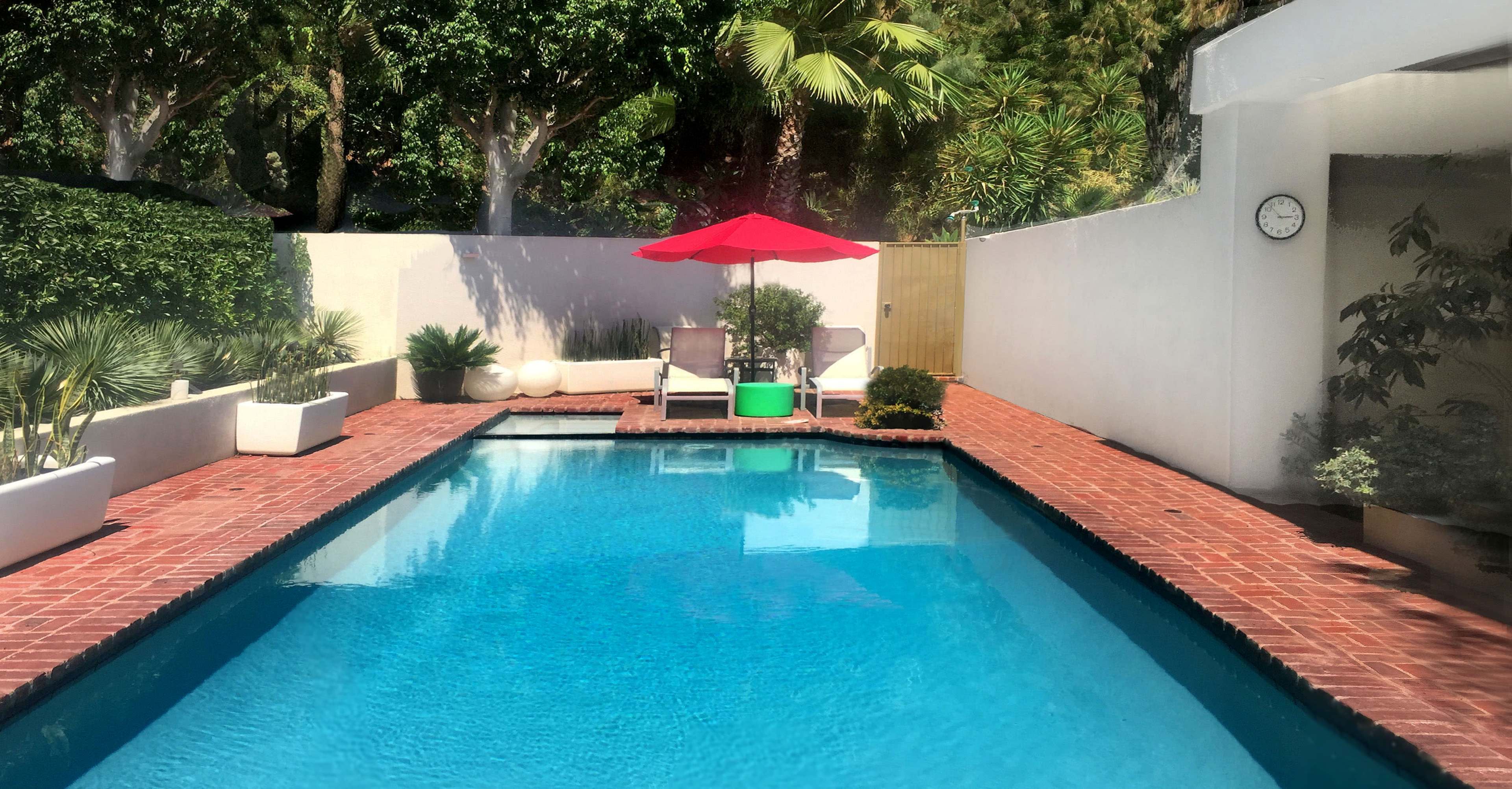 The image shows a swimming pool surrounded by greenery, with a red umbrella over a lounge chair and clock mounted on the wall.