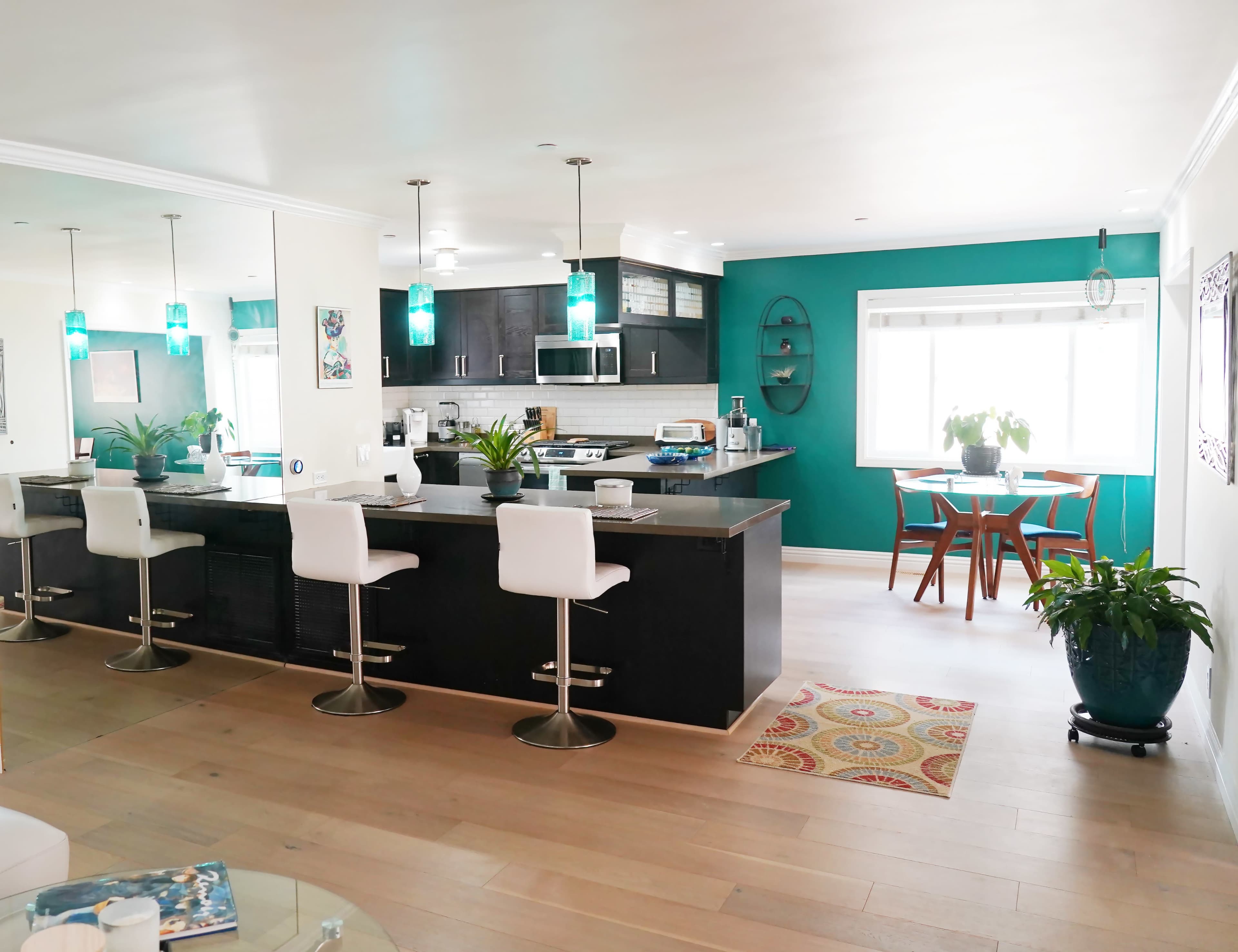 A modern kitchen with dark cabinetry and a breakfast bar, featuring teal accents, plants, and a dining area with a round table and chairs.