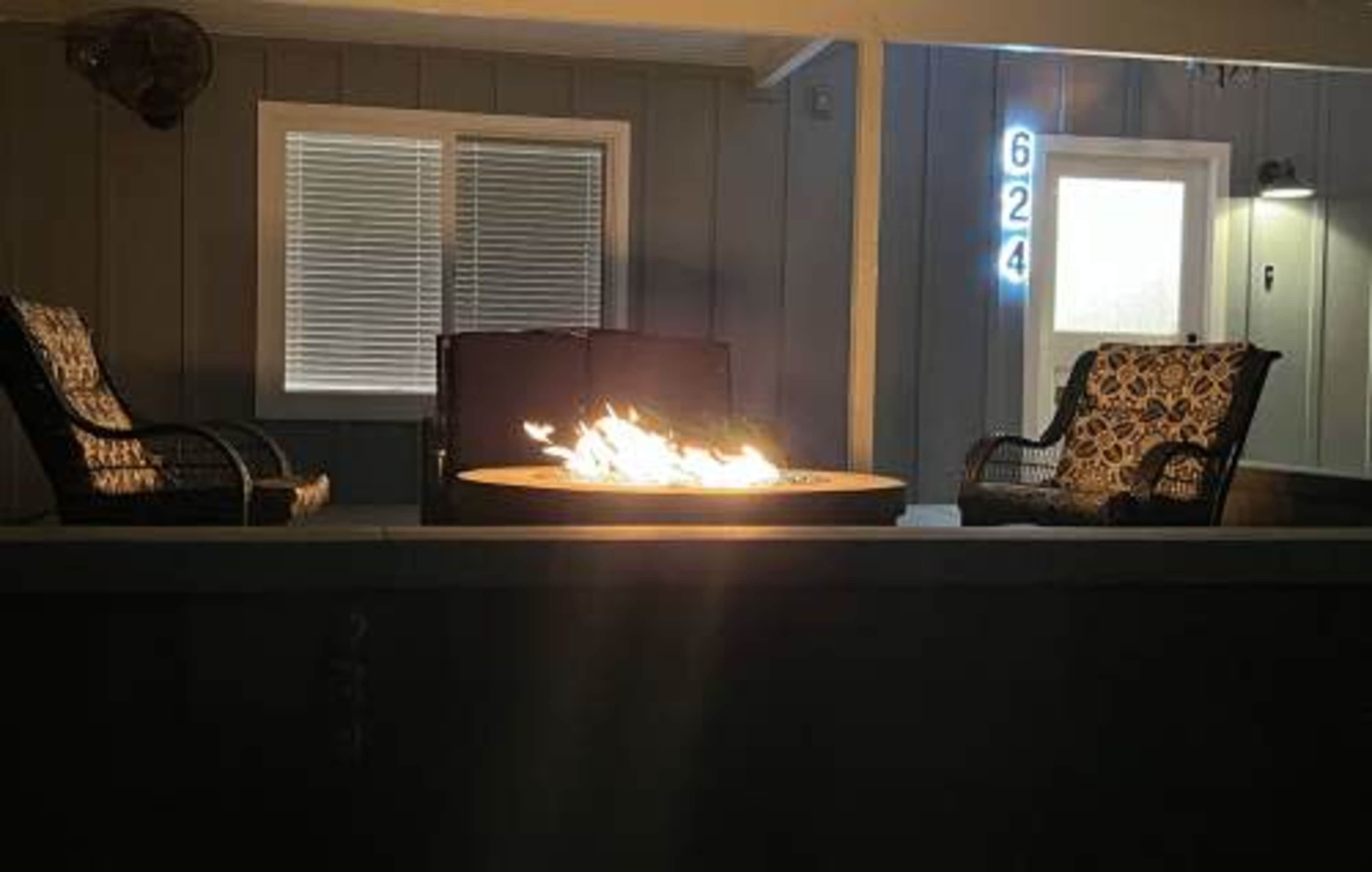 A cozy outdoor seating area features two chairs facing a fire pit with flames, illuminated by nearby lighting.