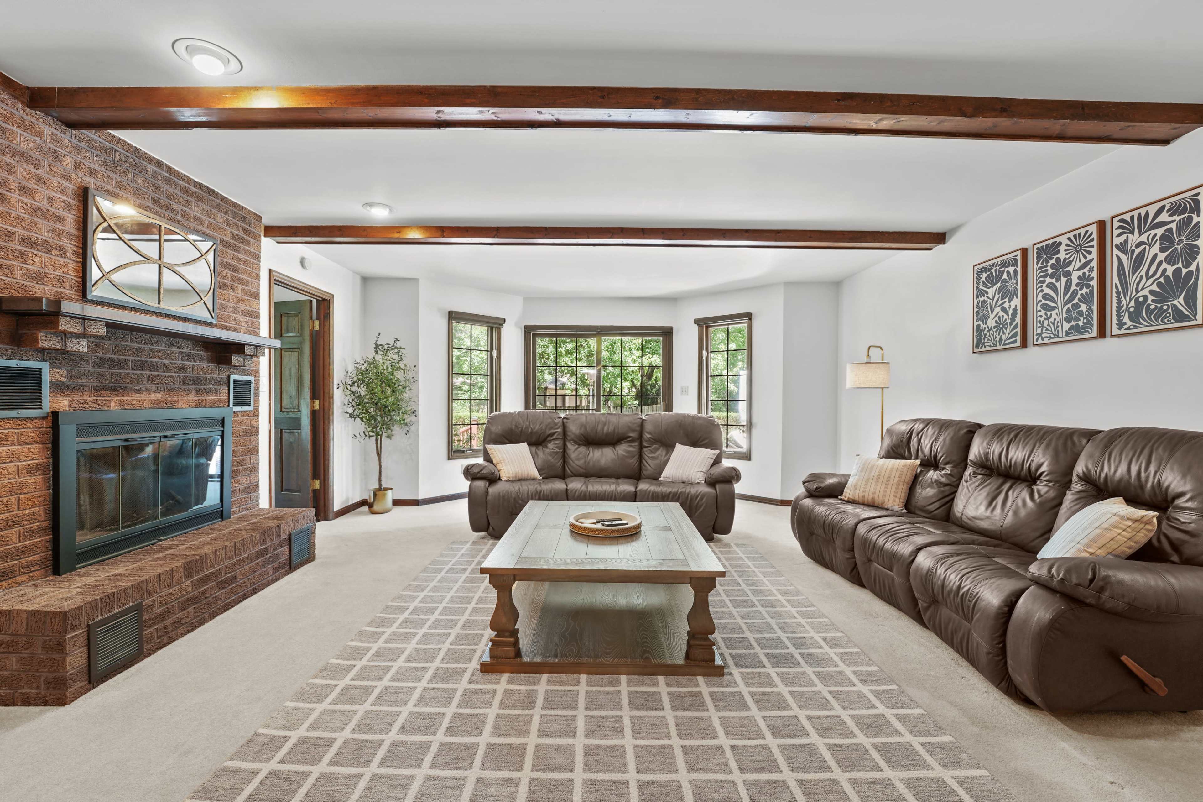 The image shows a spacious living room featuring a brown brick fireplace, two leather sofas, a wooden coffee table, and large windows with natural light.