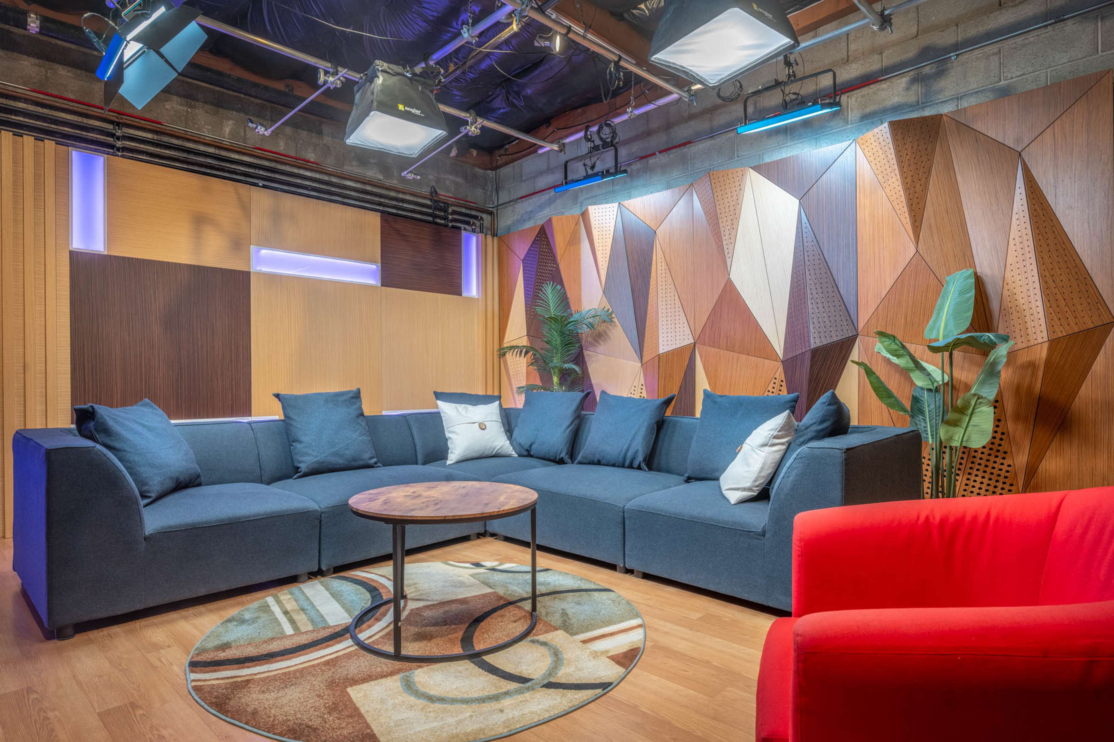 A modern lounge area featuring a blue sectional sofa, a round wooden coffee table, and a red accent chair, all set against a backdrop of geometric wooden panels and soft lighting.