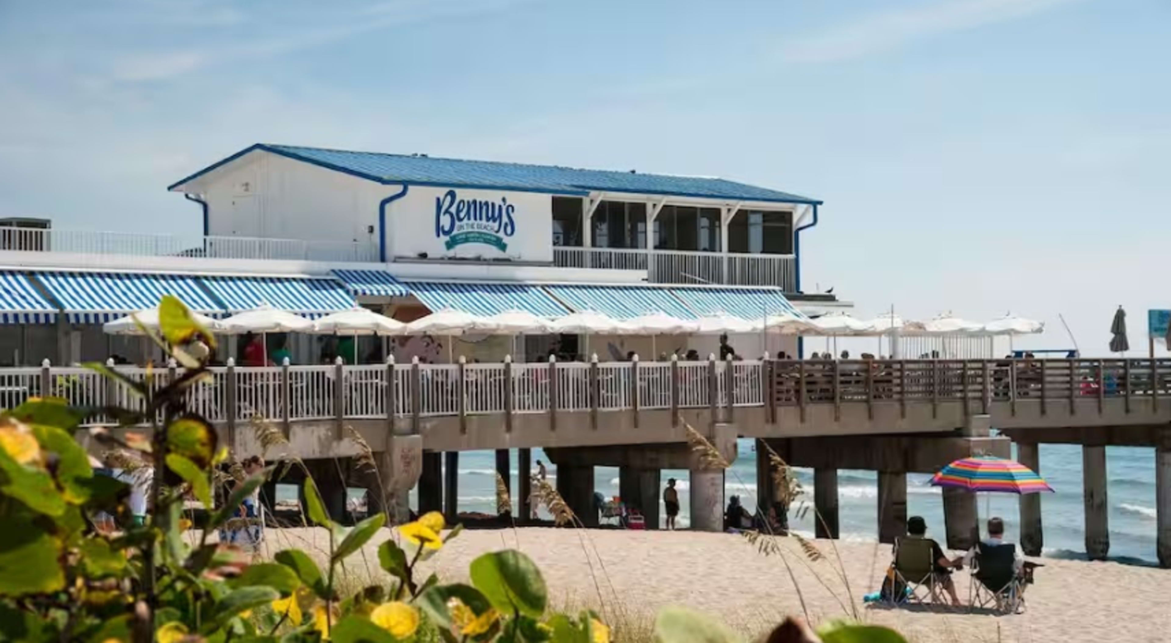 The image shows a beachfront restaurant named Benny's, positioned on a pier with people sitting under umbrellas on the sandy beach.