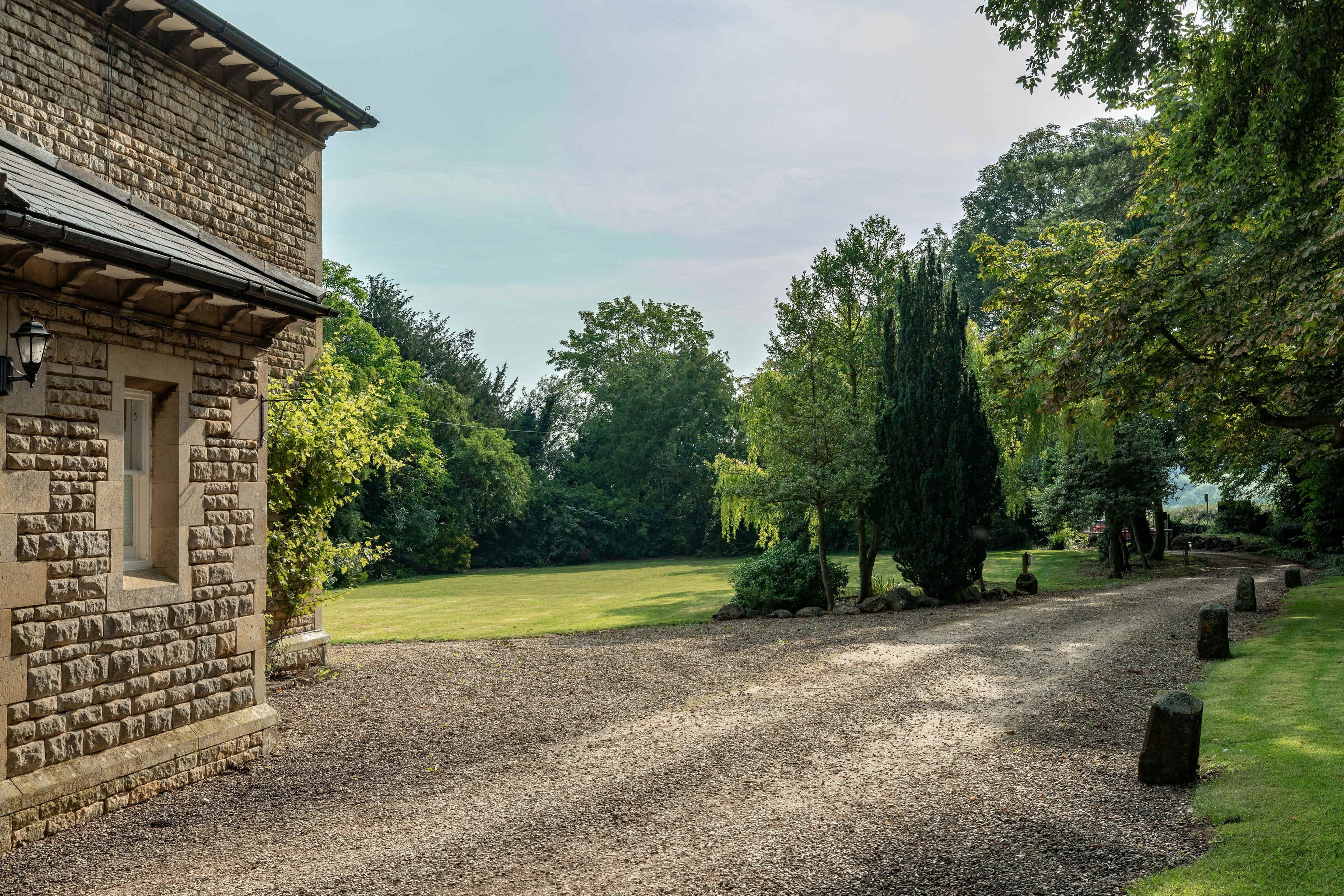 A gravel pathway leads alongside a stone building, bordered by trees and a spacious green lawn.