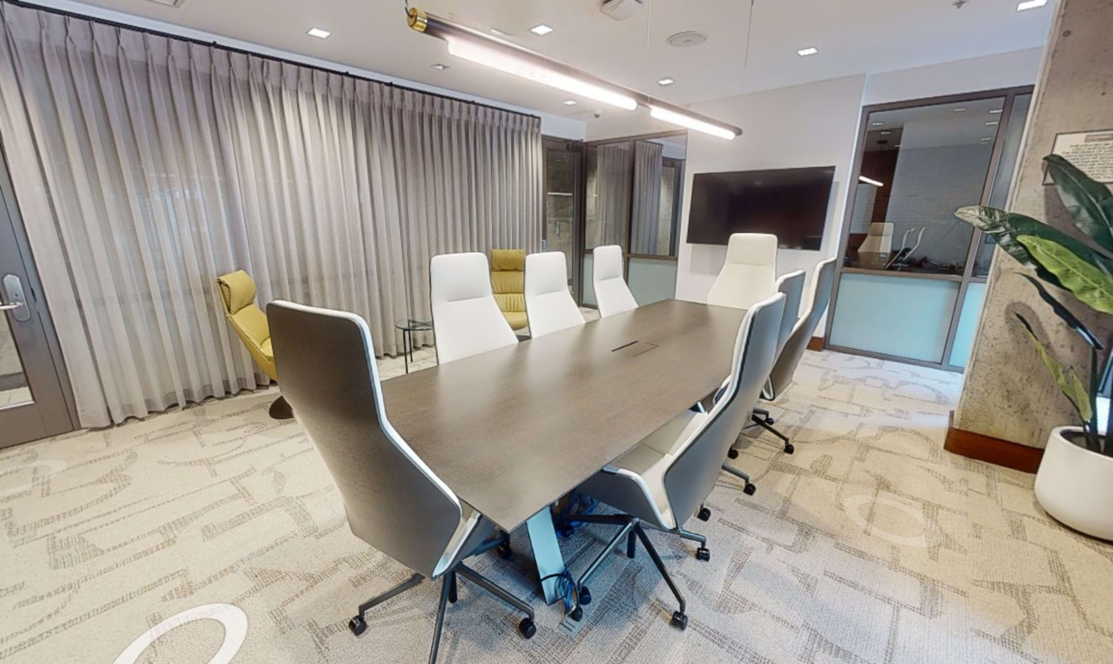 A conference room with a long wooden table surrounded by white ergonomic chairs, a large screen on the wall, and floor-to-ceiling curtains.
