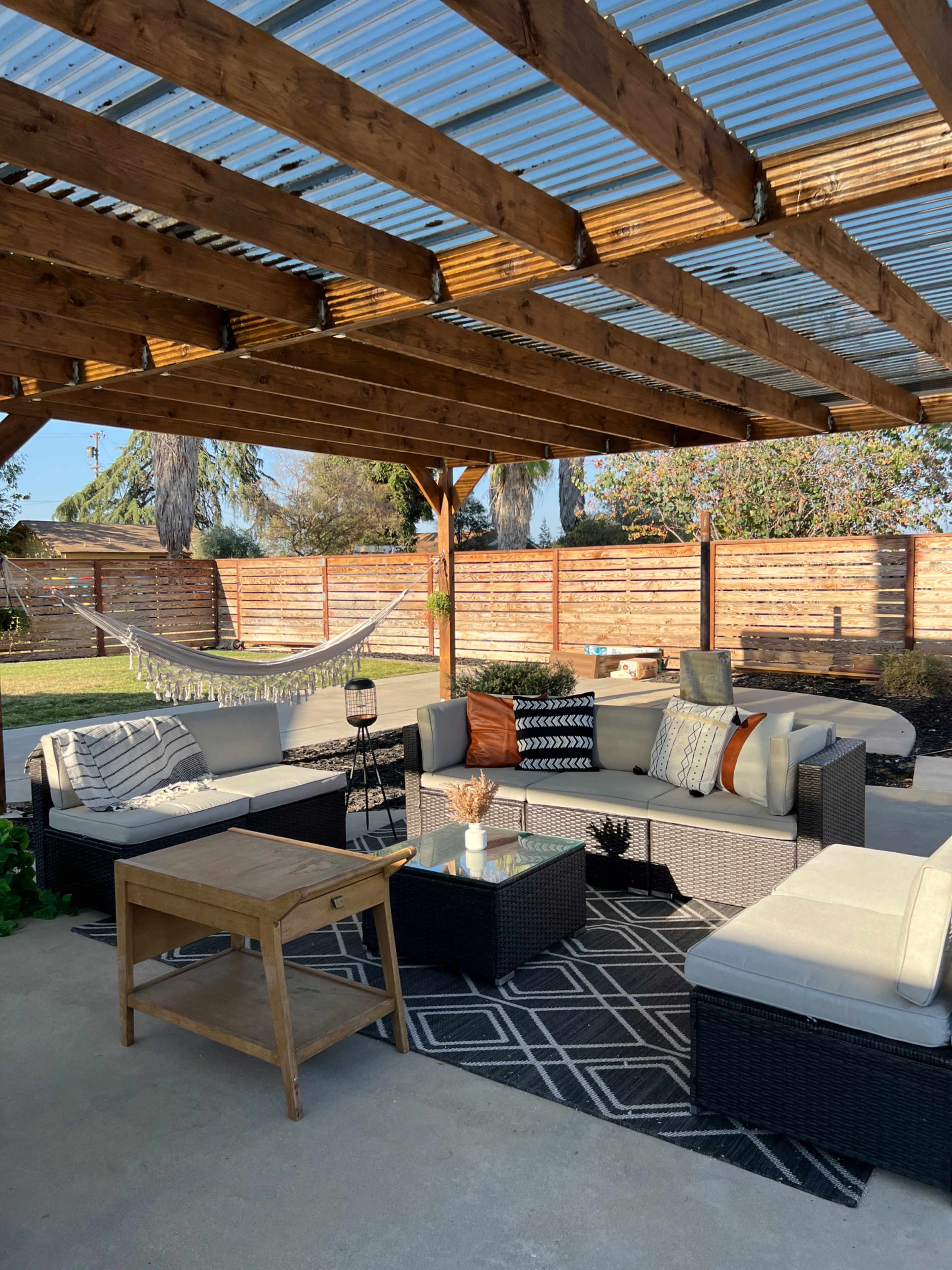 An outdoor patio is arranged with a sectional sofa, coffee table, and a hammock beneath a wooden pergola.