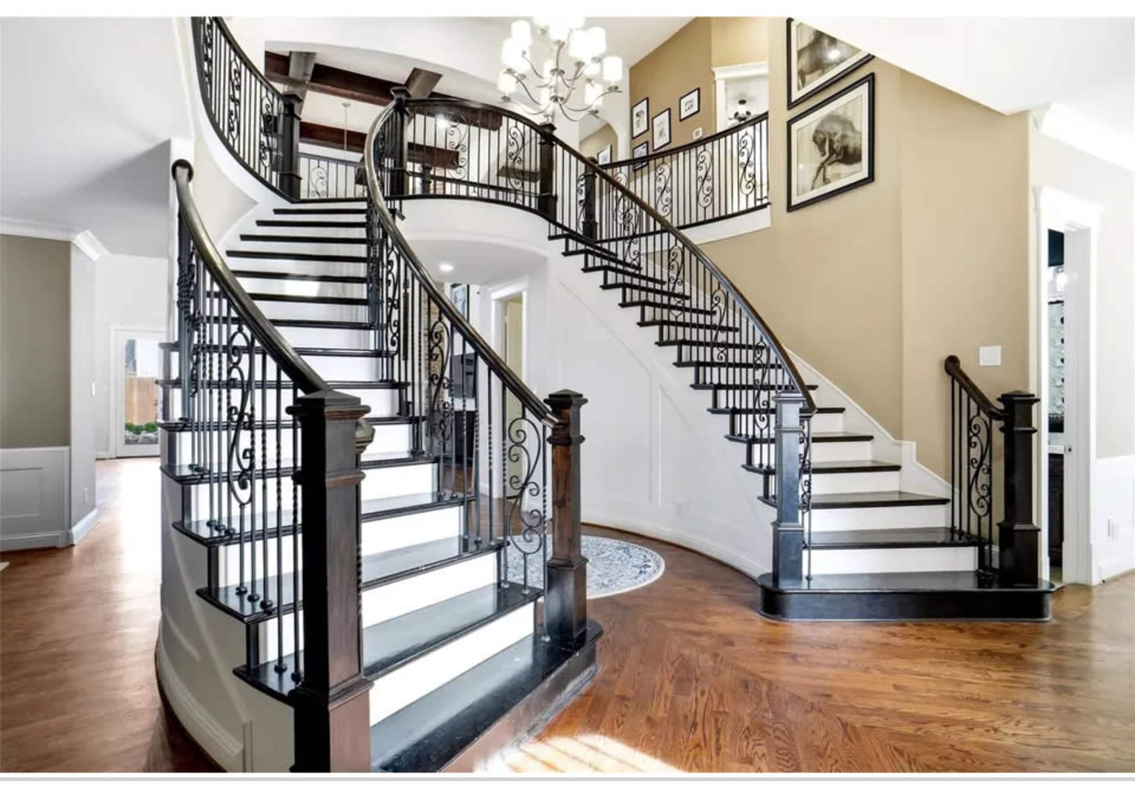 The image shows a grand, curved staircase with black and white steps and decorative wrought iron railings, situated in an elegantly designed foyer.