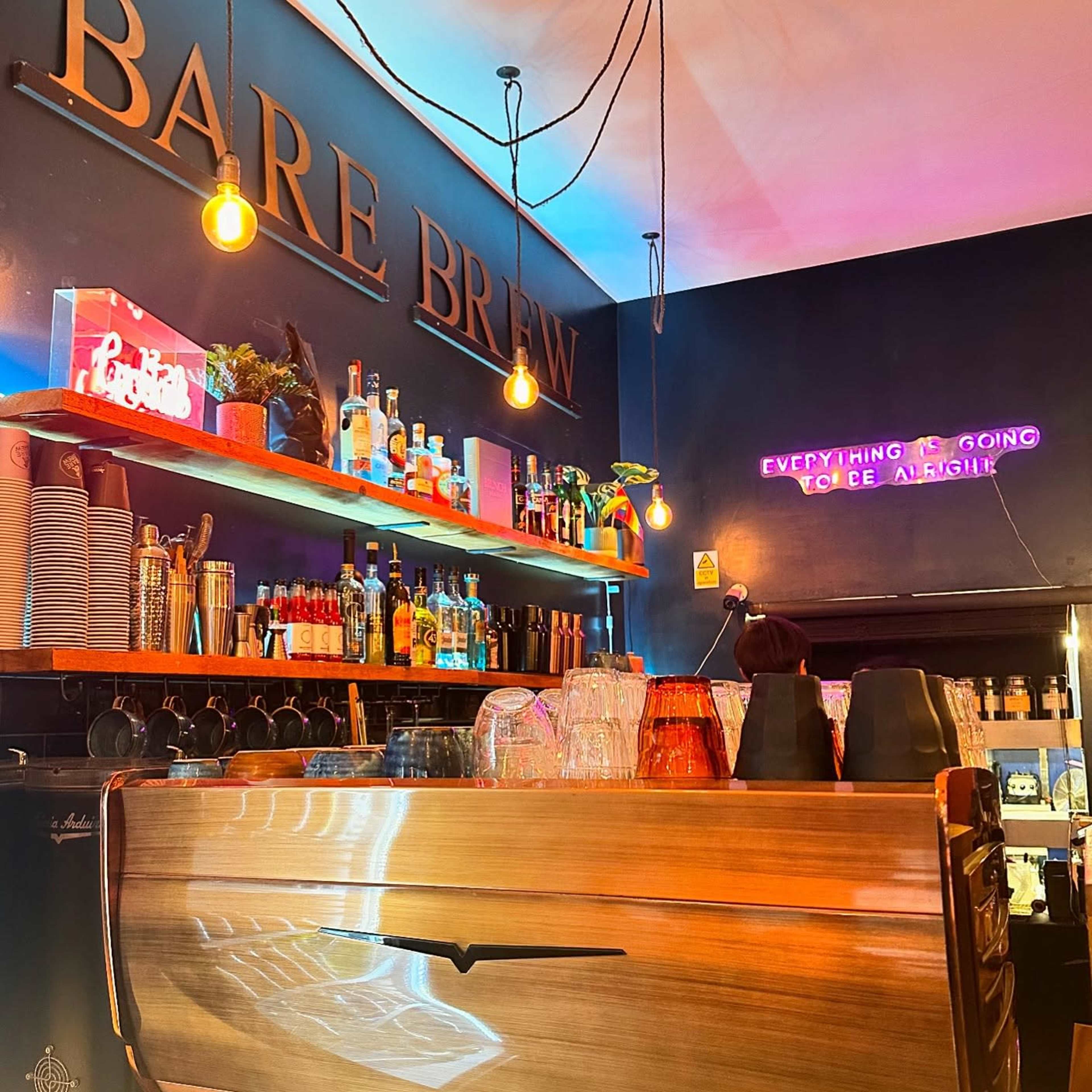 A brightly lit coffee shop interior features a polished wooden counter with various bottles and cups displayed on shelves above, complemented by a neon sign that states, "everything is going coffee alright."