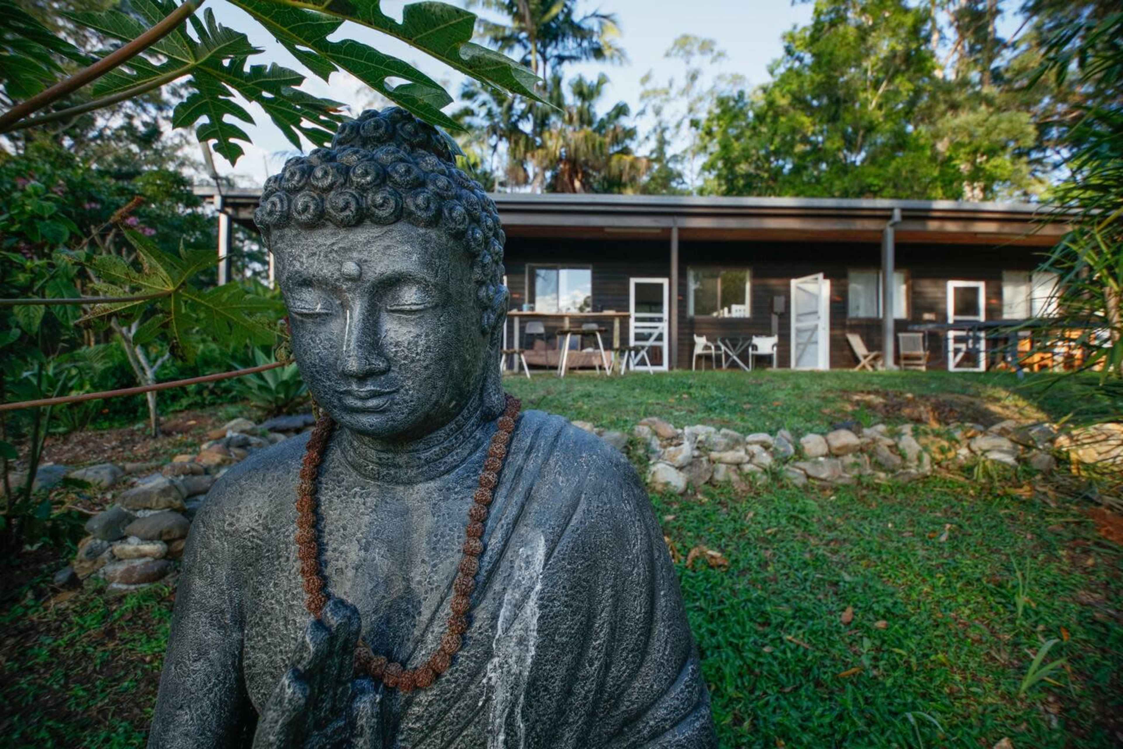 A stone Buddha statue is positioned in a garden with a wooden building and outdoor seating in the background.