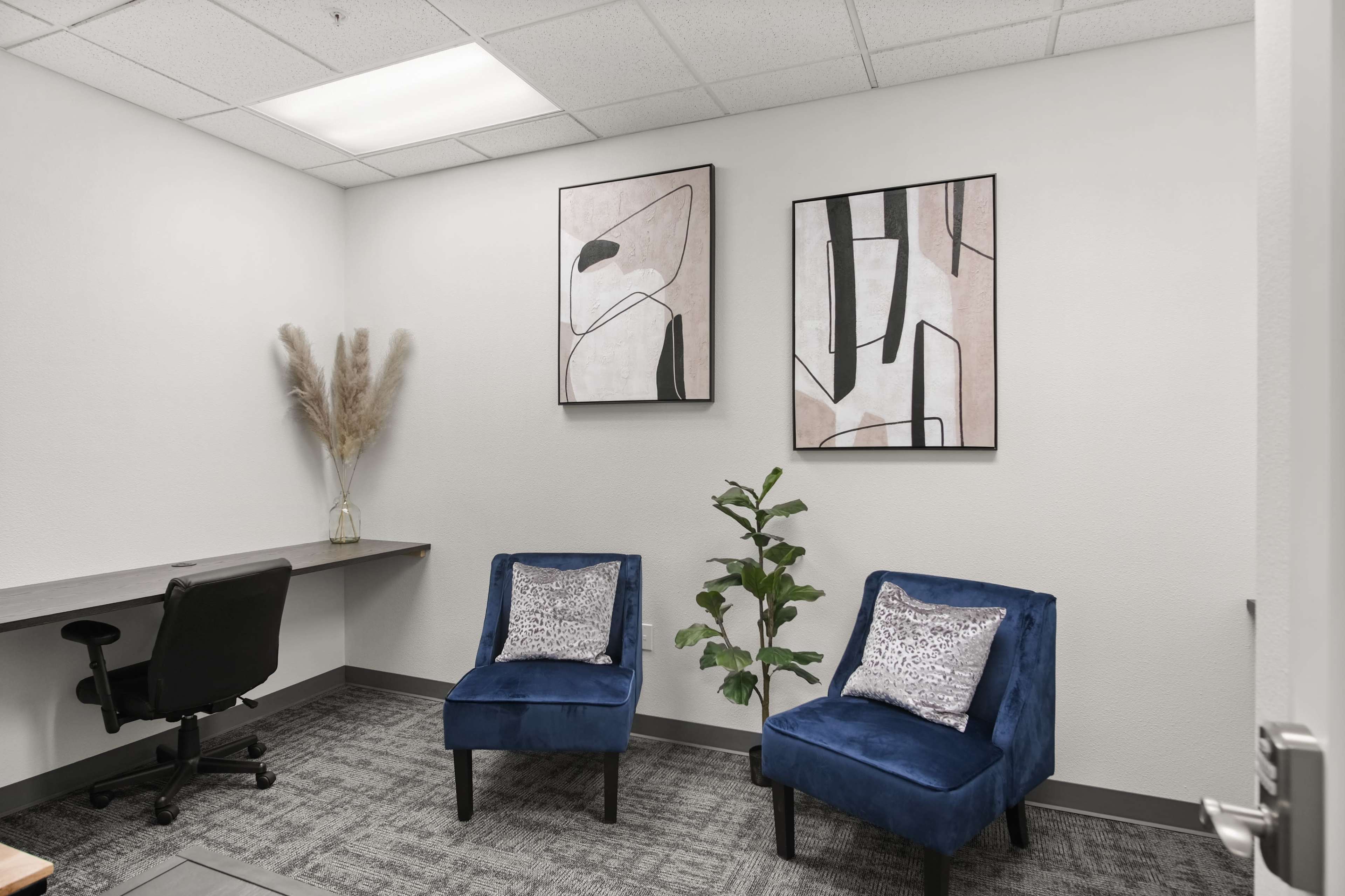 A minimalist office space with two blue chairs, a small plant, a desk with a chair, and two abstract framed artworks on the walls.