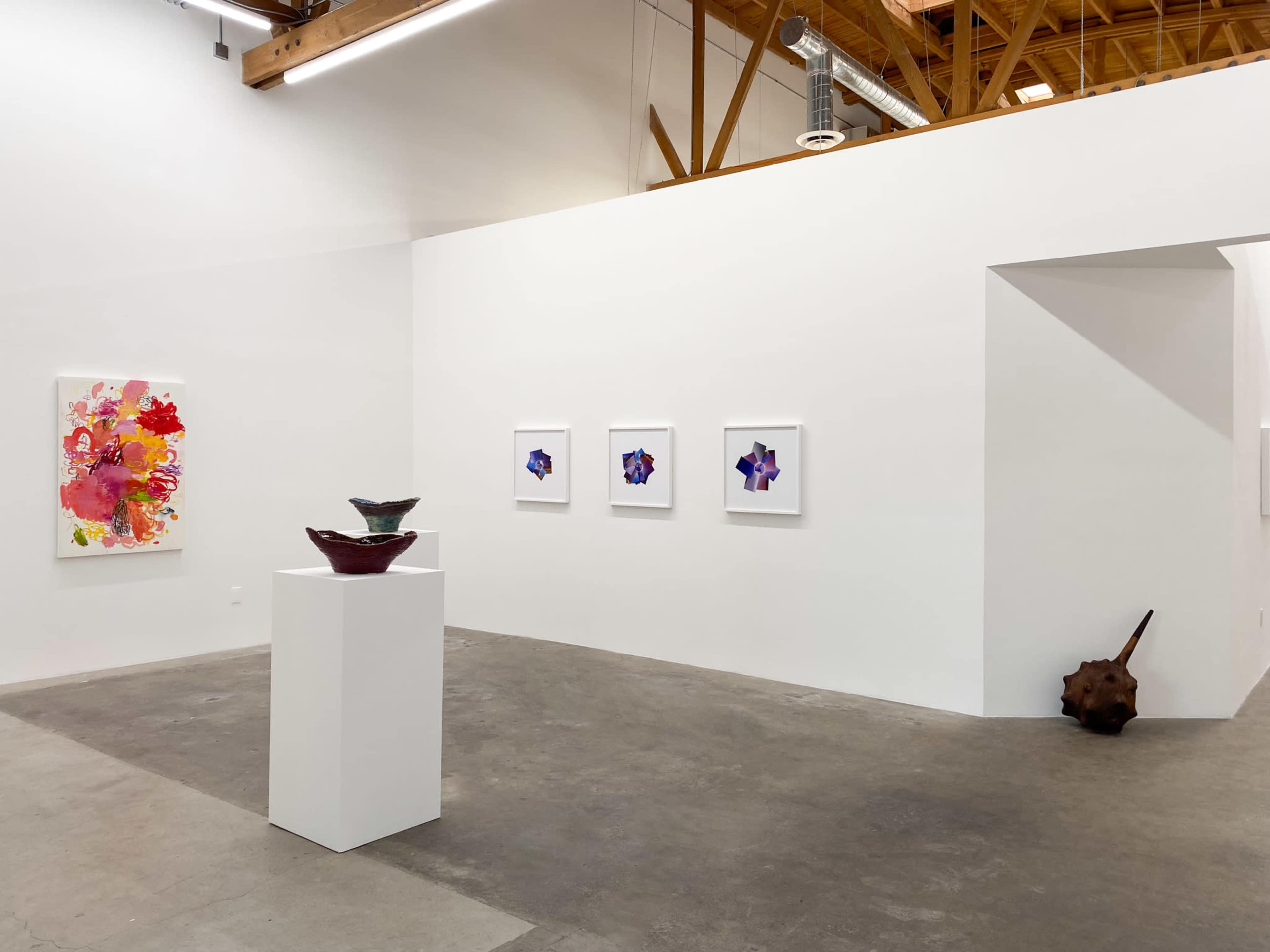 An art gallery featuring a white wall with several framed artworks and a pedestal display with two bowls on it.