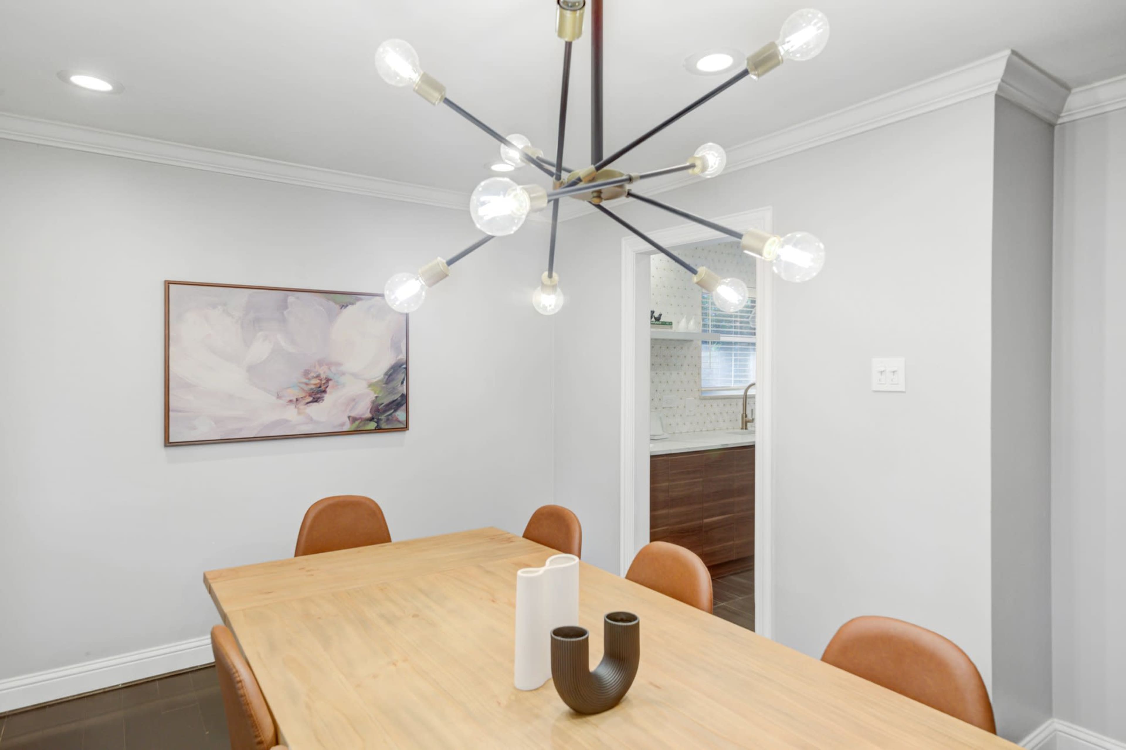 A modern dining room features a wooden table surrounded by brown chairs, with a unique light fixture and a floral artwork on the wall.