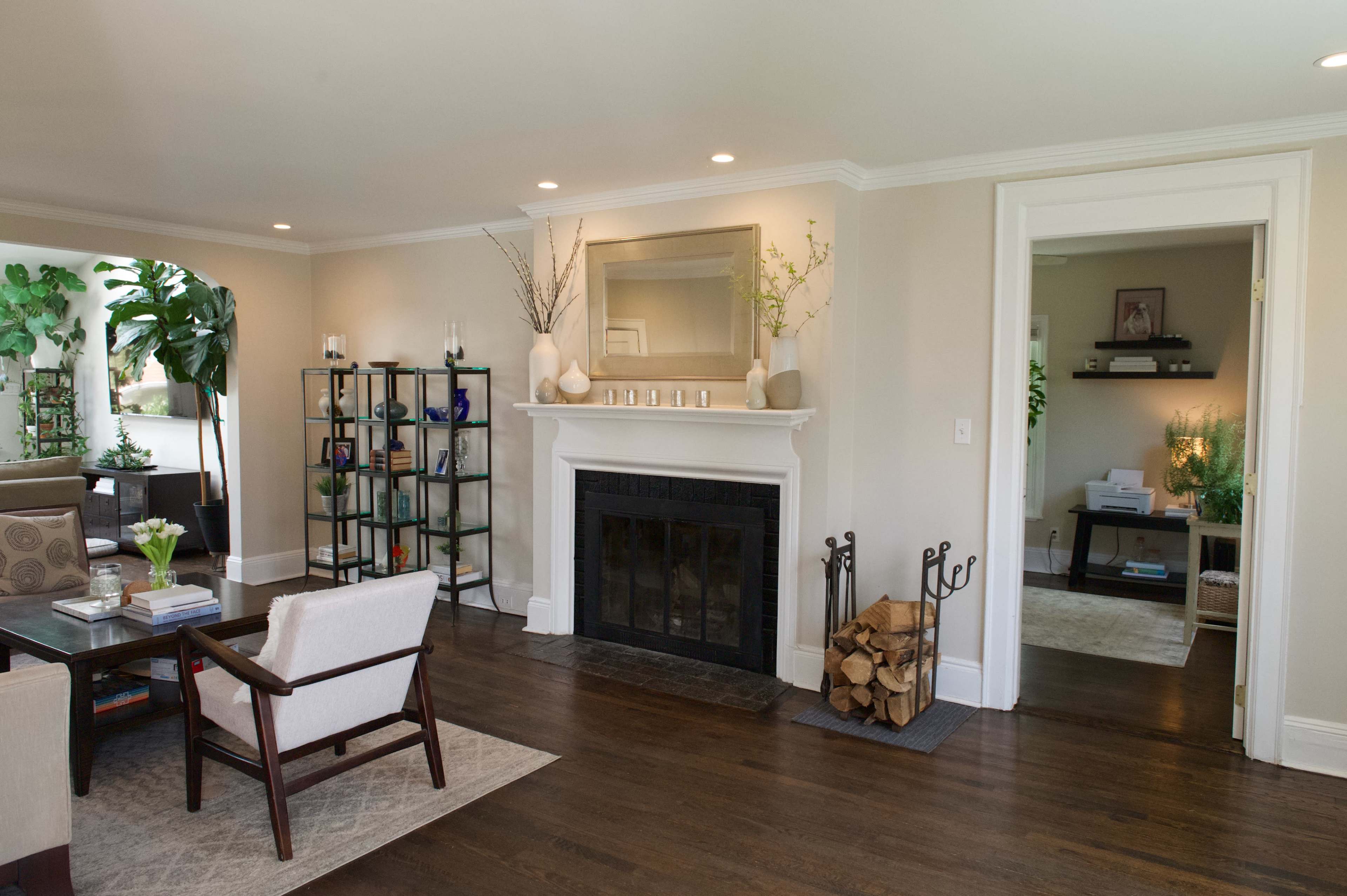 The image shows a modern living room featuring a fireplace, a bookshelf, and a seating area with a coffee table, complemented by decorative plants and a doorway leading to another room.