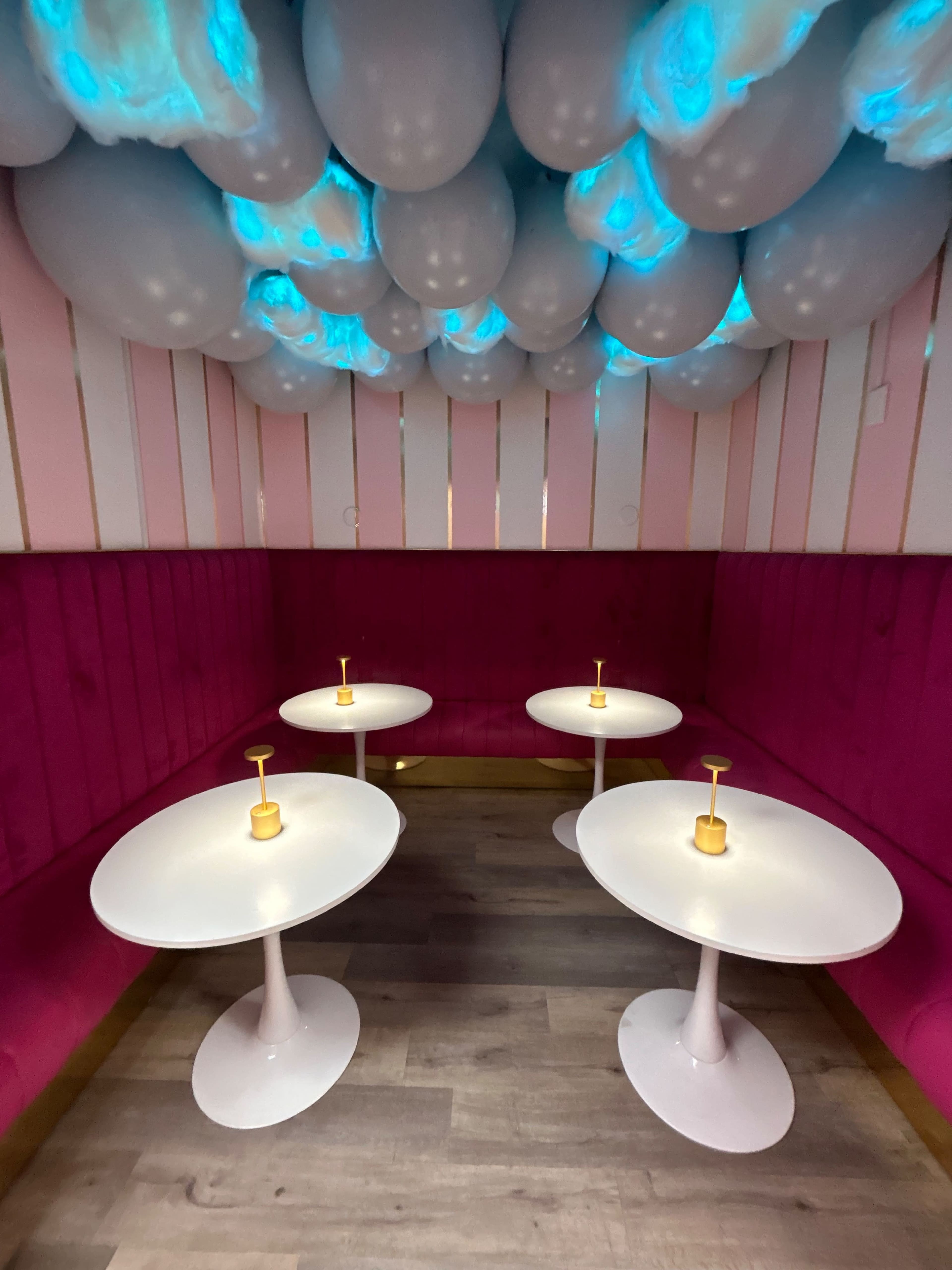 A cozy seating area features four white tables with yellow candle holders, under a ceiling adorned with large gray balloons that emit soft blue light.