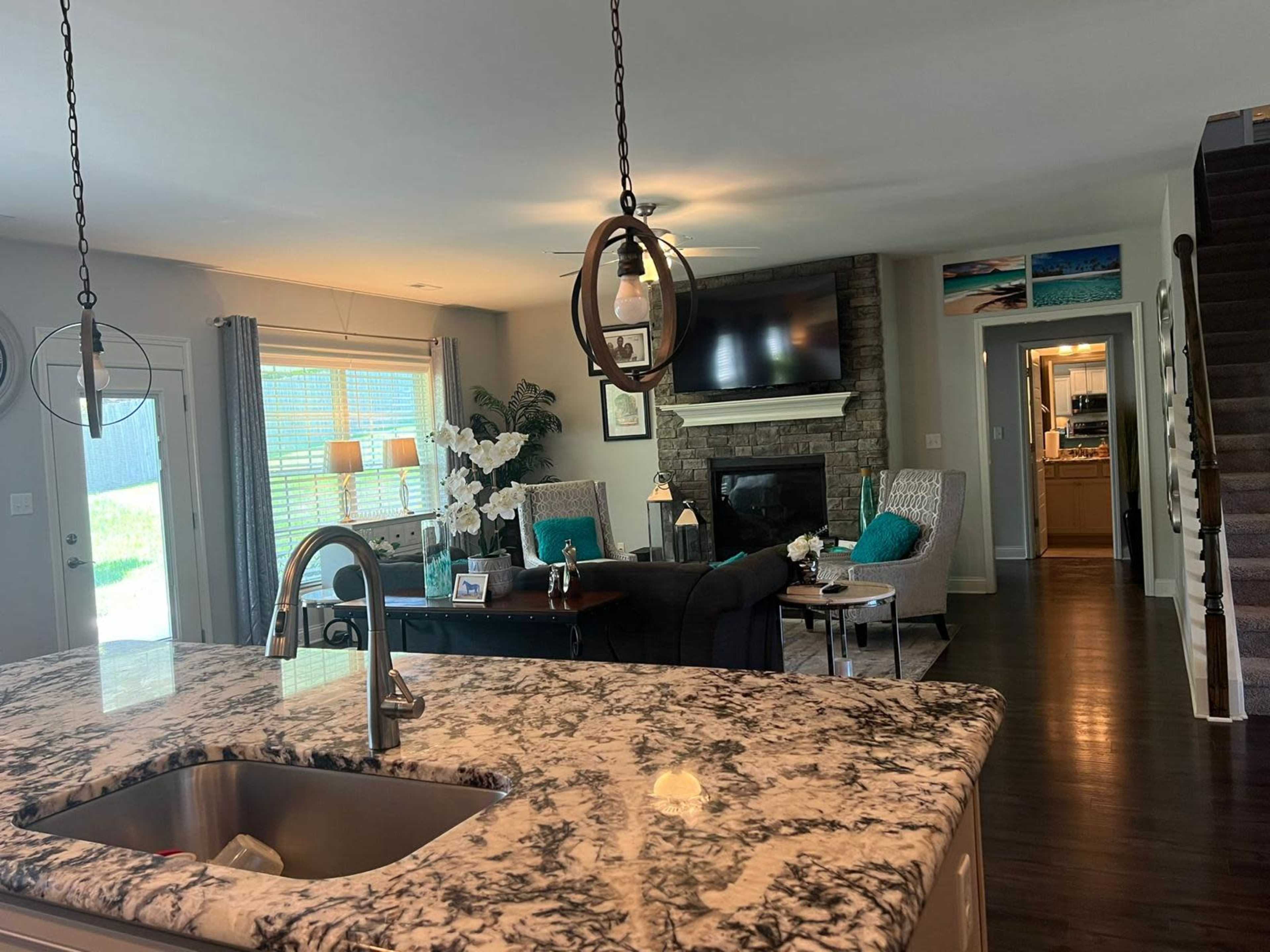 A living room with a stone fireplace and large windows, viewed from a kitchen with a granite countertop.