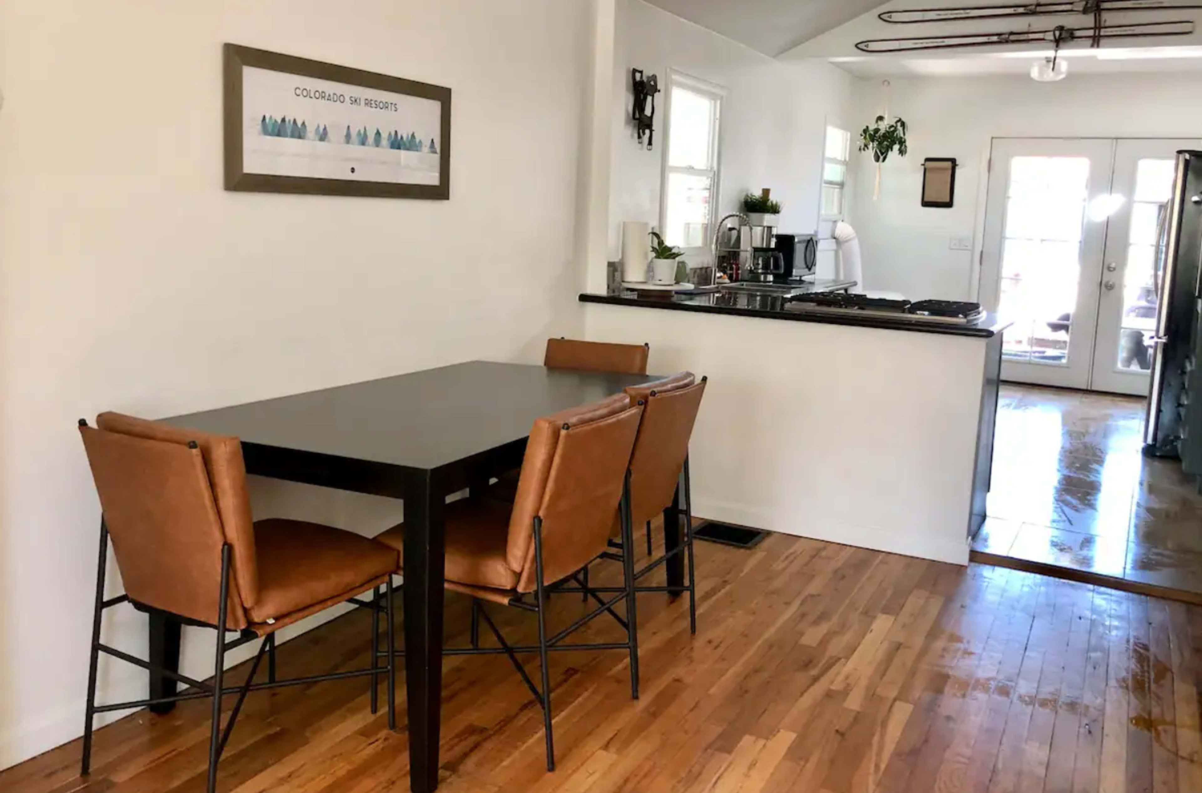 A simple dining area features a rectangular table with four brown chairs, adjacent to a kitchen with modern appliances and a doorway leading outside.