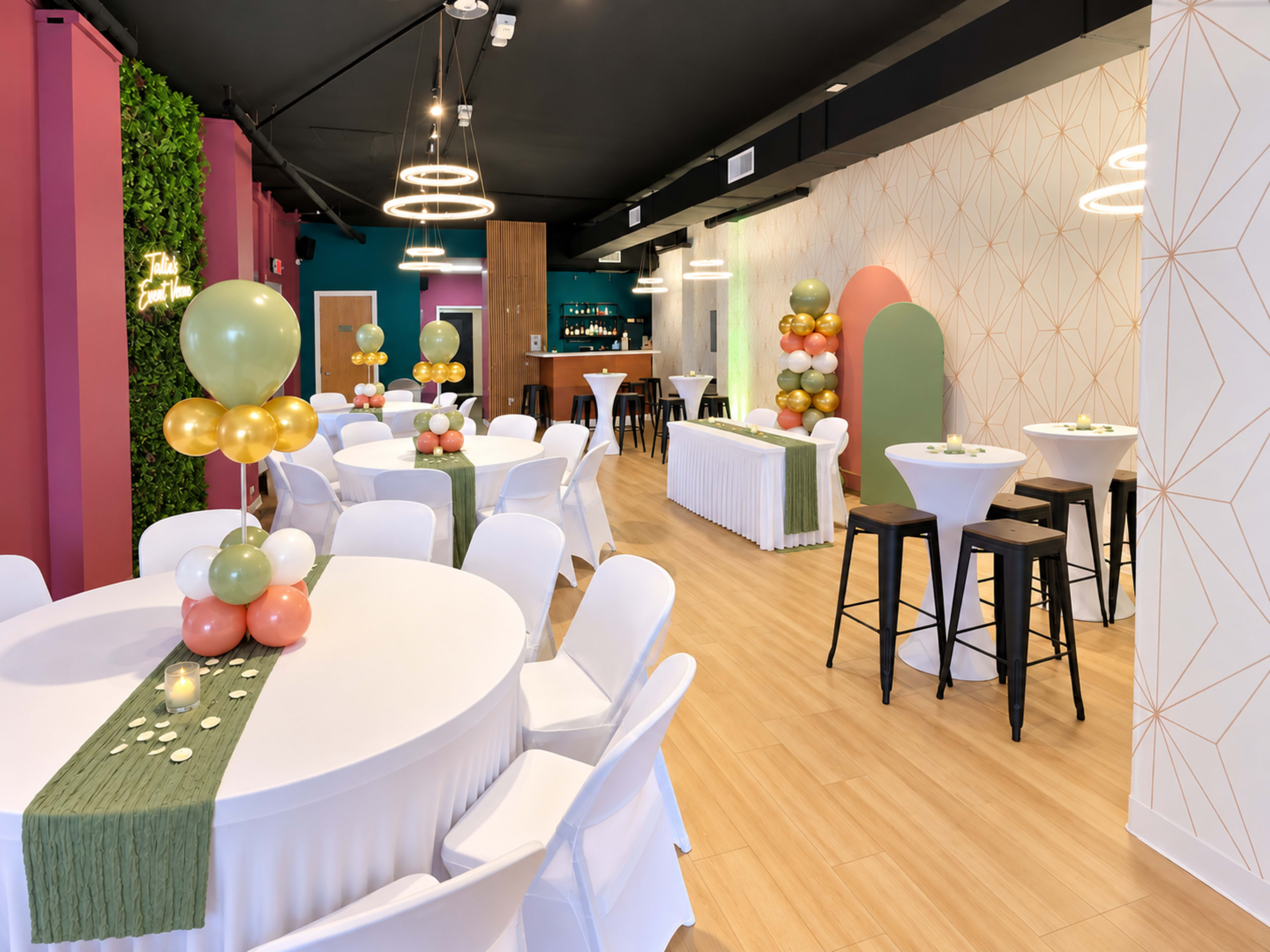 The image shows a modern event space decorated with round tables covered in white tablecloths, colorful balloon arrangements, and a bar area in the background.