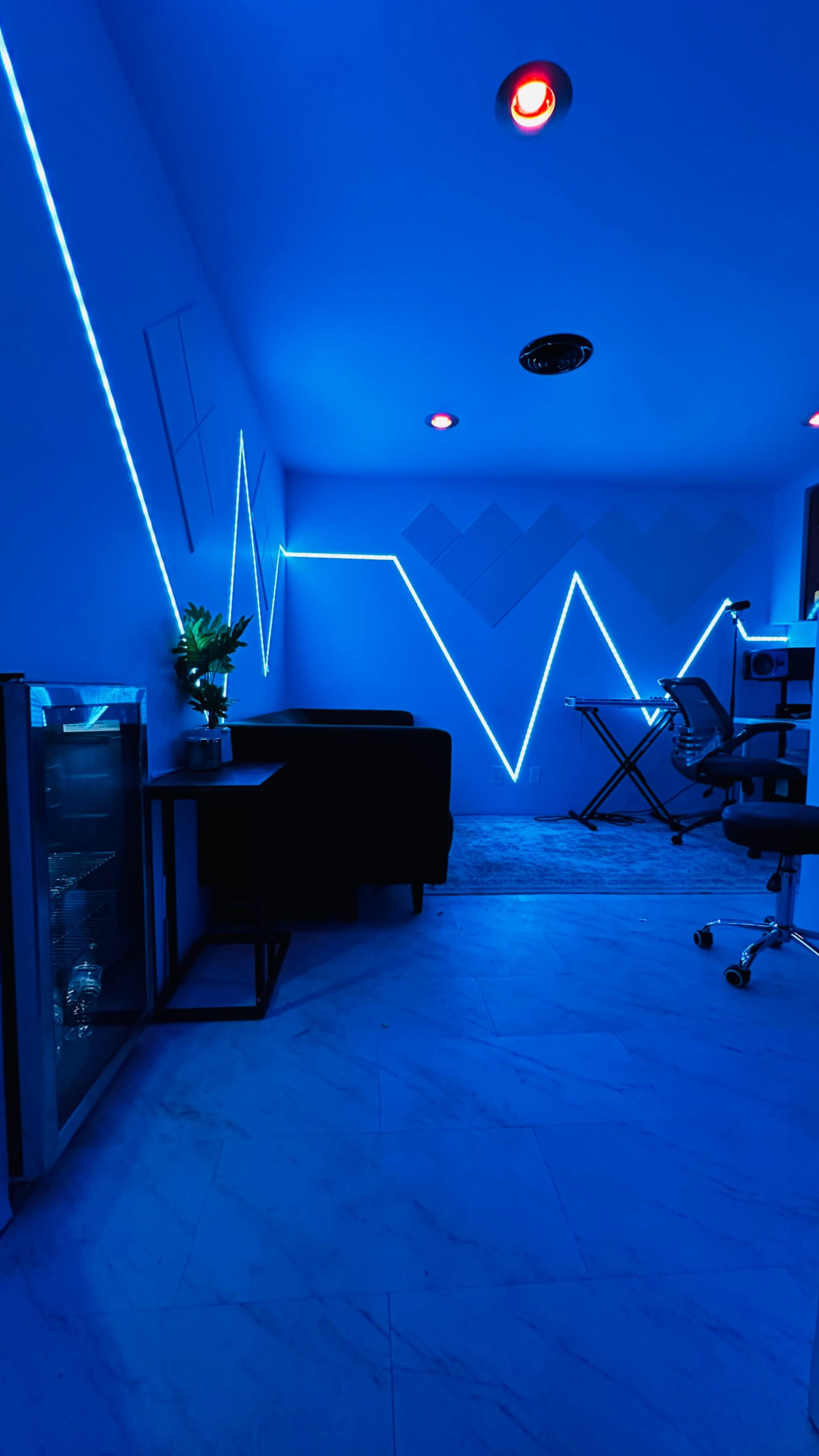 A modern room features blue LED light lines on the walls, a couch, a small table, a chair, and a desk with equipment.