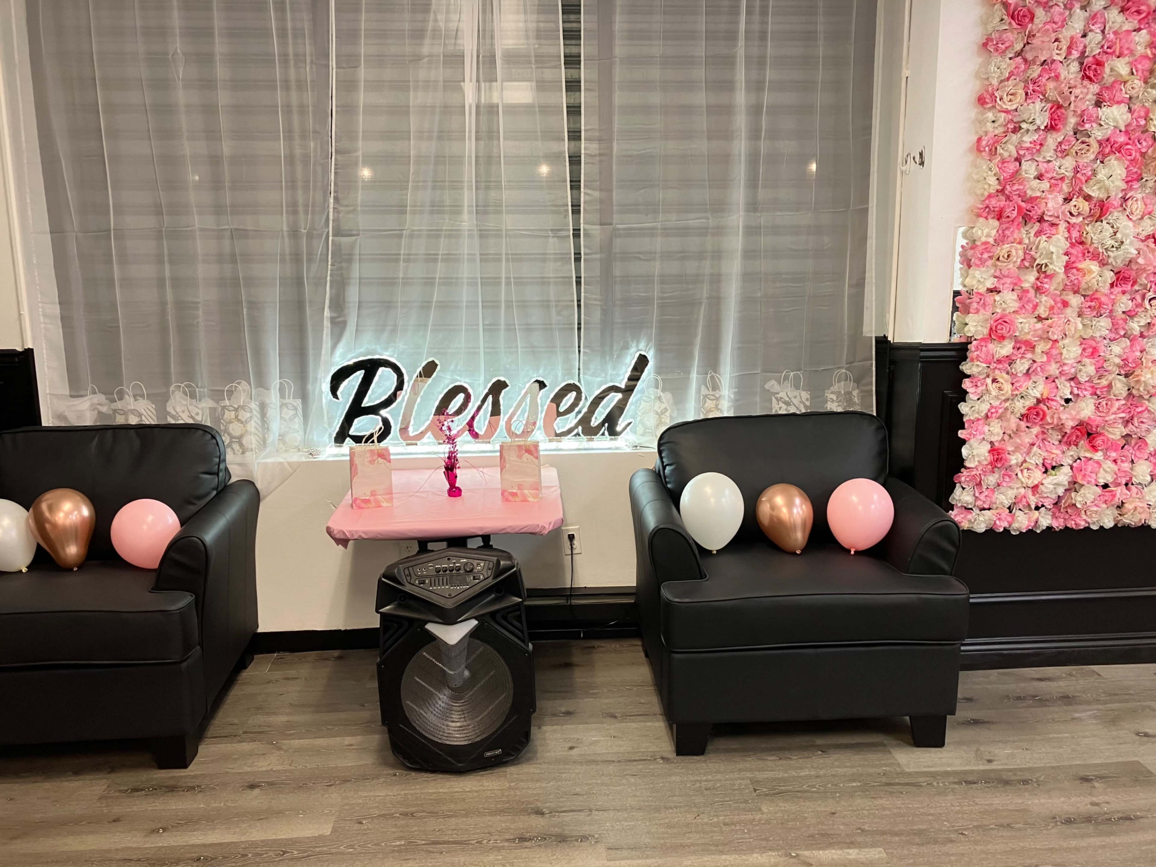 The scene features two black leather chairs with balloons and a pink table underneath a sign that reads "Blessed," alongside a wall decorated with pink flowers.