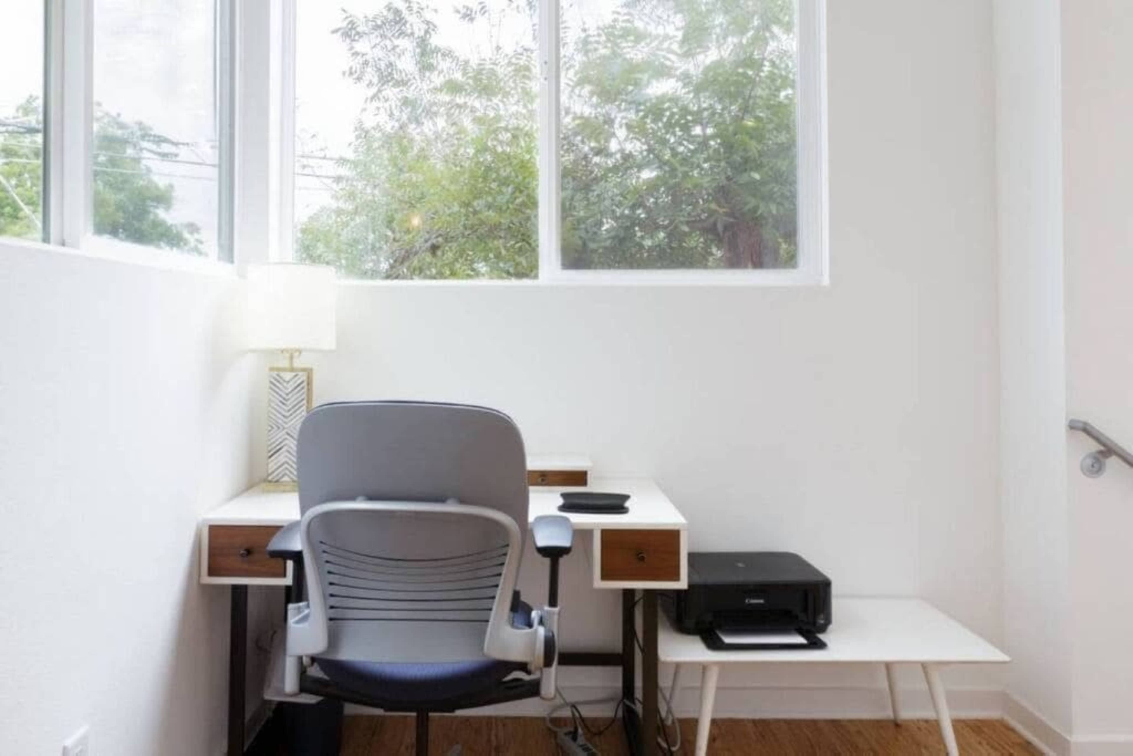 A modern workspace features a desk with a chair, a lamp, and a printer, all situated near a large window with greenery outside.