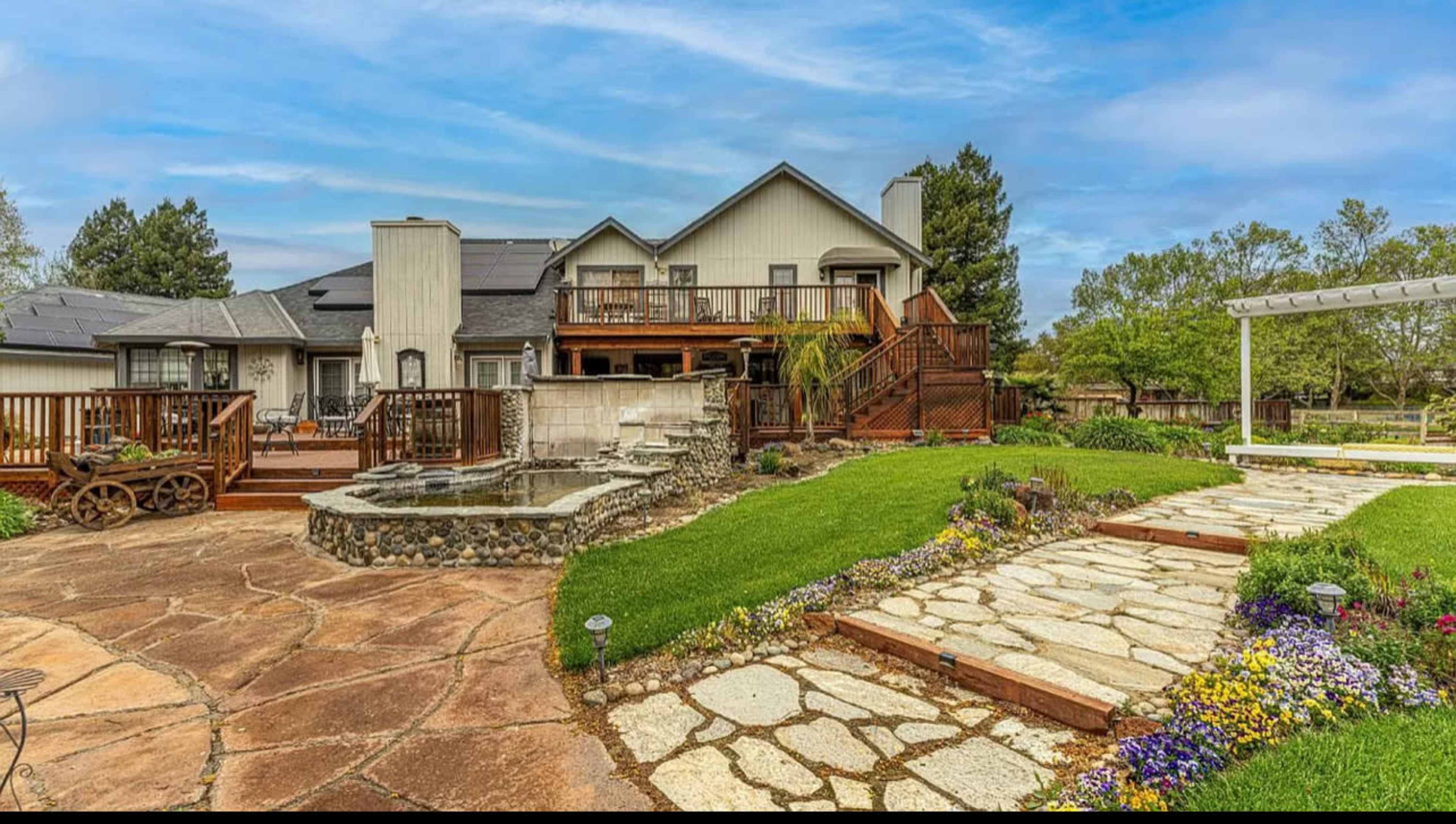 A landscaped backyard with a stone path leading to a decked house, a water feature, and flower beds.