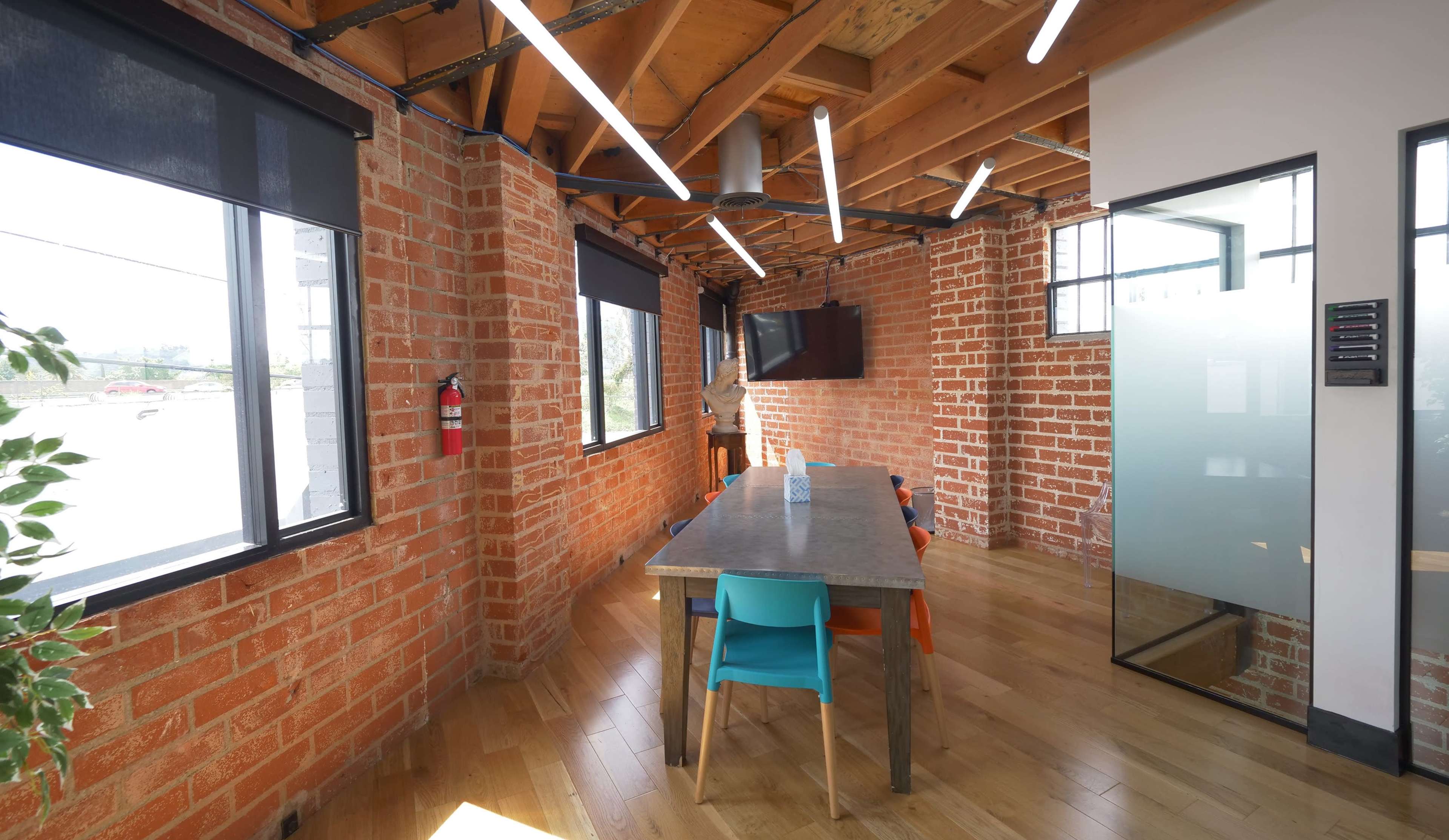 The image shows a modern office space with exposed brick walls, a wooden table surrounded by colorful chairs, and large windows allowing natural light to enter.