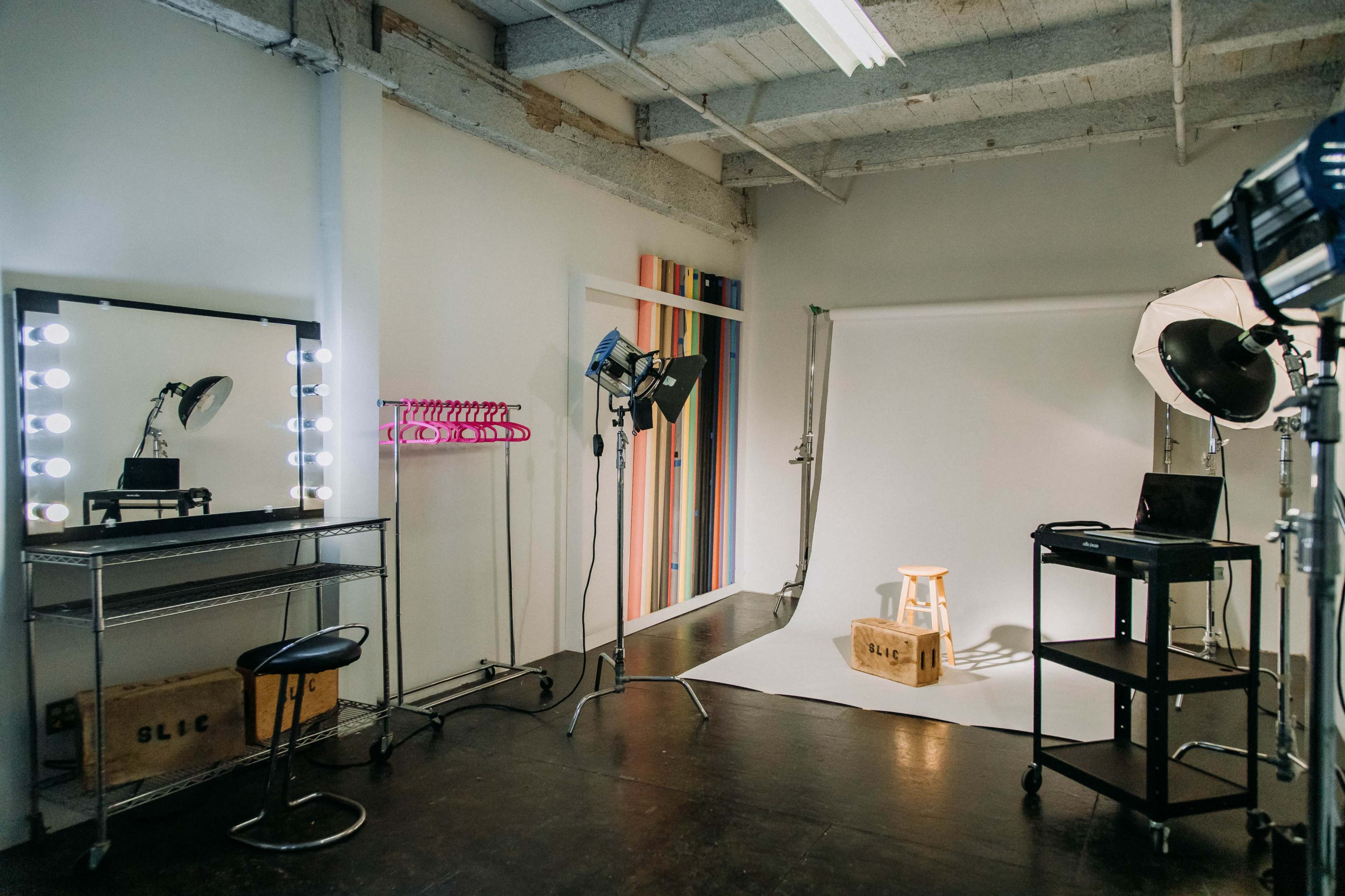 A photography studio featuring a white backdrop, a stool, lighting equipment, and a mirrored vanity station.