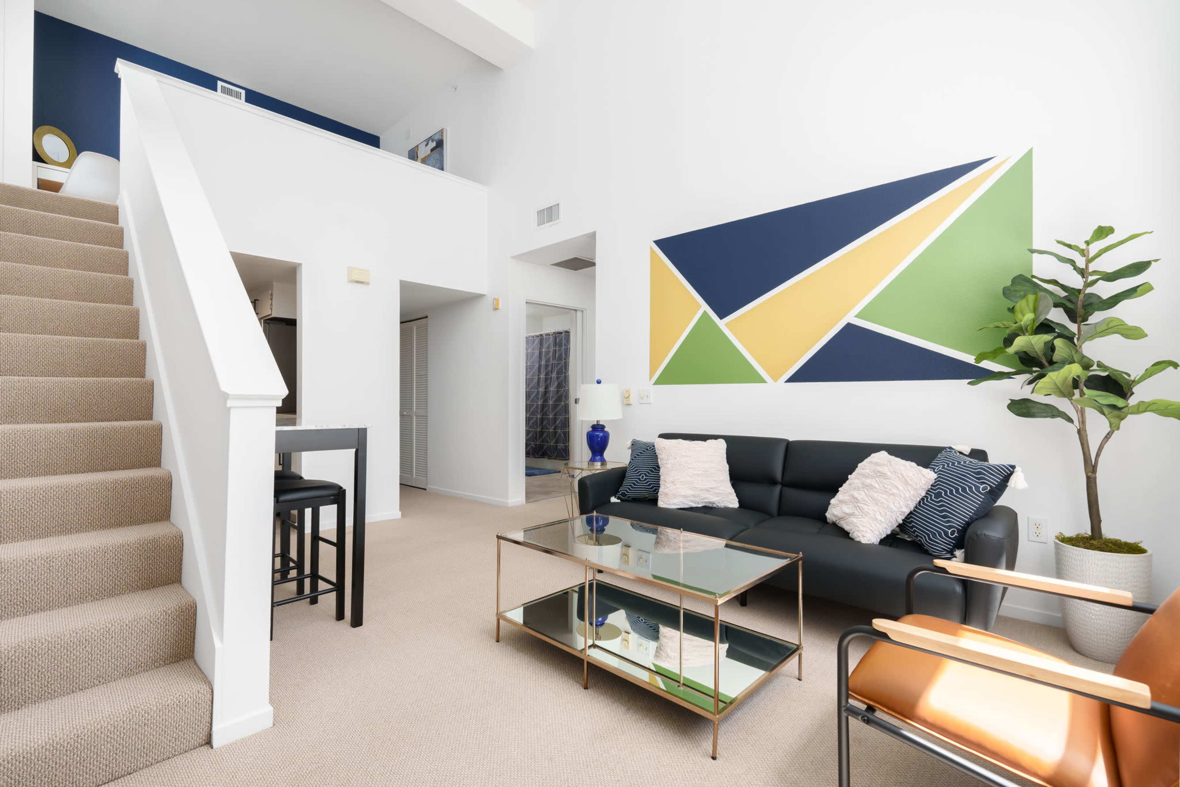 A modern living room featuring a black sofa, a glass coffee table, a potted plant, and a colorful geometric wall mural, with a staircase visible on the left.