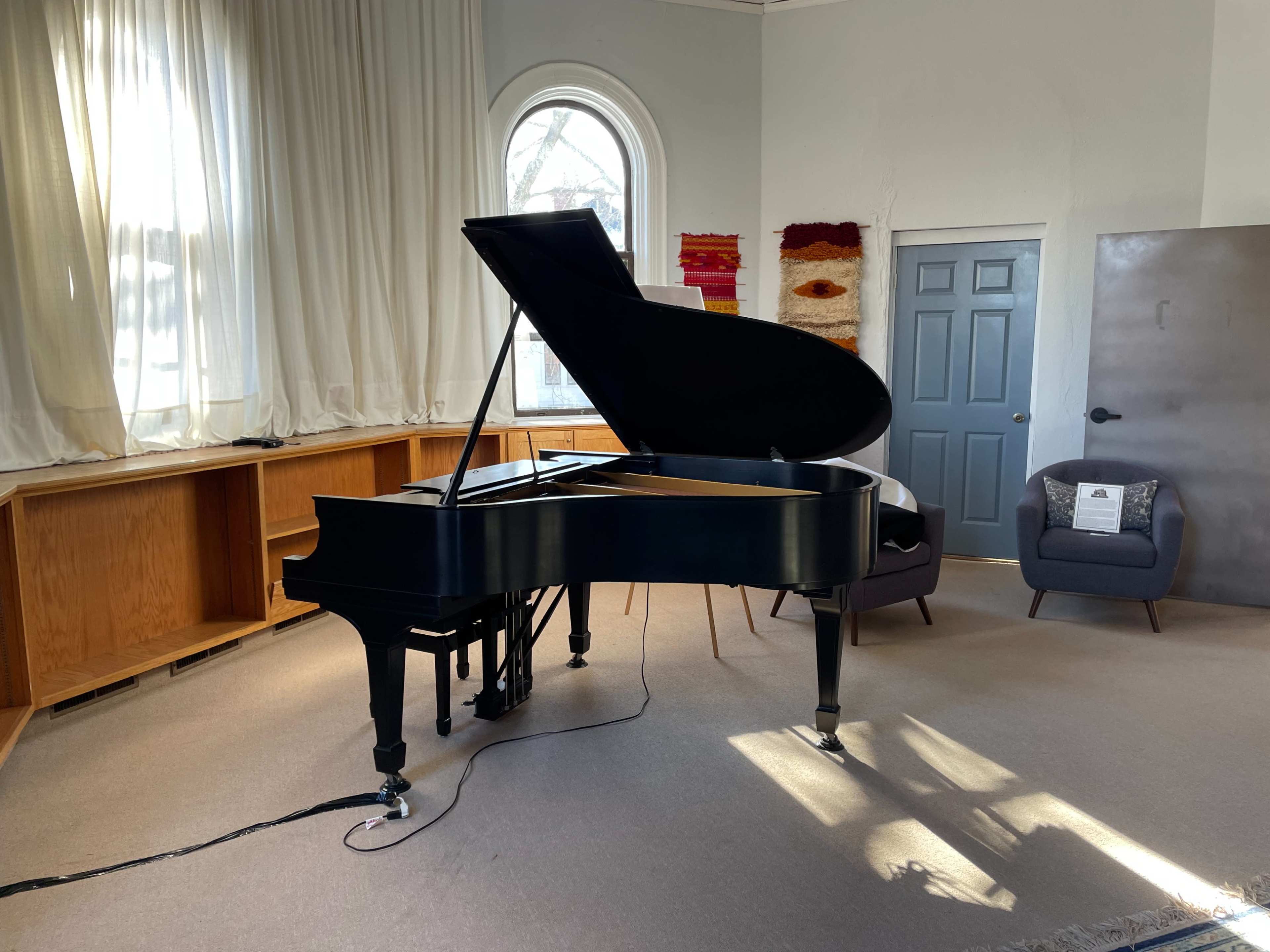 A black grand piano stands in a well-lit room with large windows, surrounded by a bookshelf and a chair.