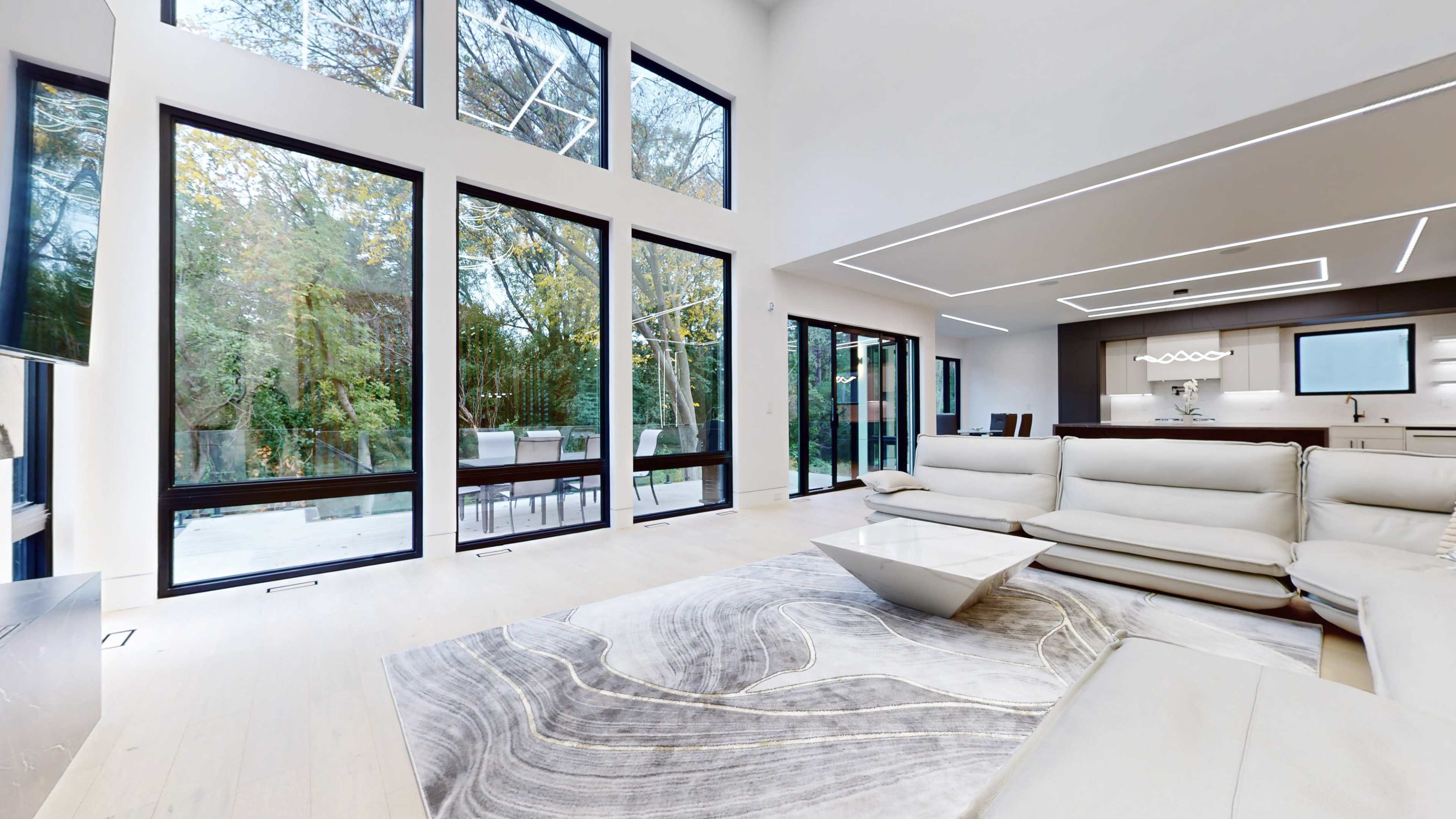 A modern living room with large floor-to-ceiling windows, light-colored furniture, and an open layout connecting to a dining area.