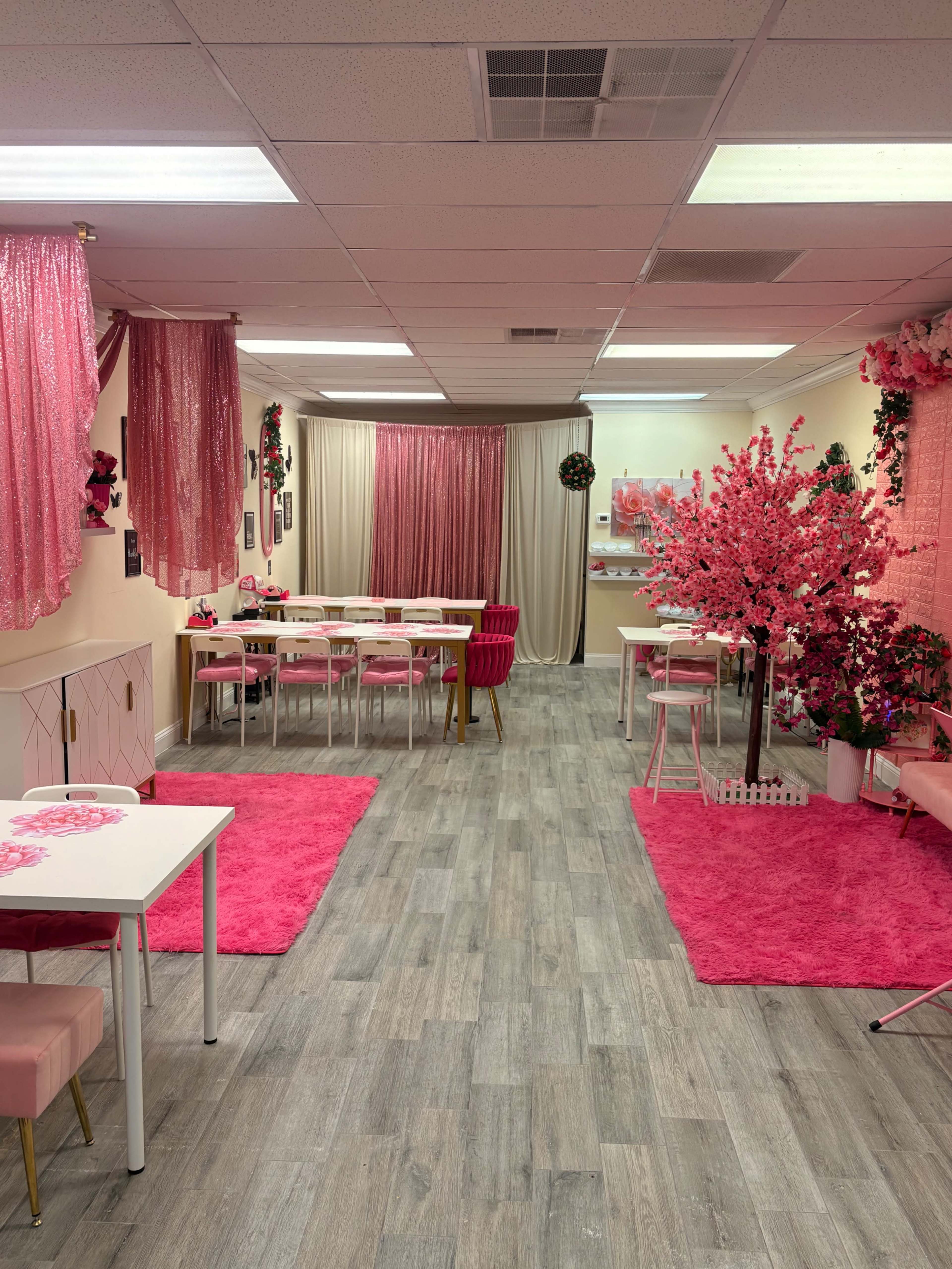 The image shows a brightly decorated room featuring pink drapes, tables with white surfaces, and a pink carpet, arranged with decorative elements like flowers and artificial trees.