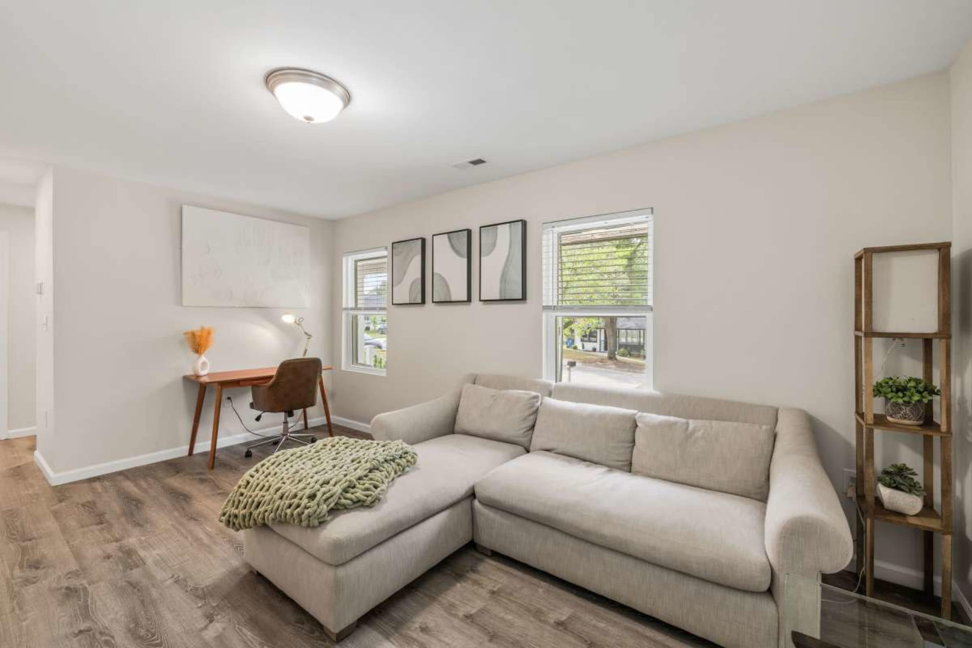The image shows a modern, well-lit living room featuring a beige sectional sofa, a desk with a lamp, and framed artwork on the walls.
